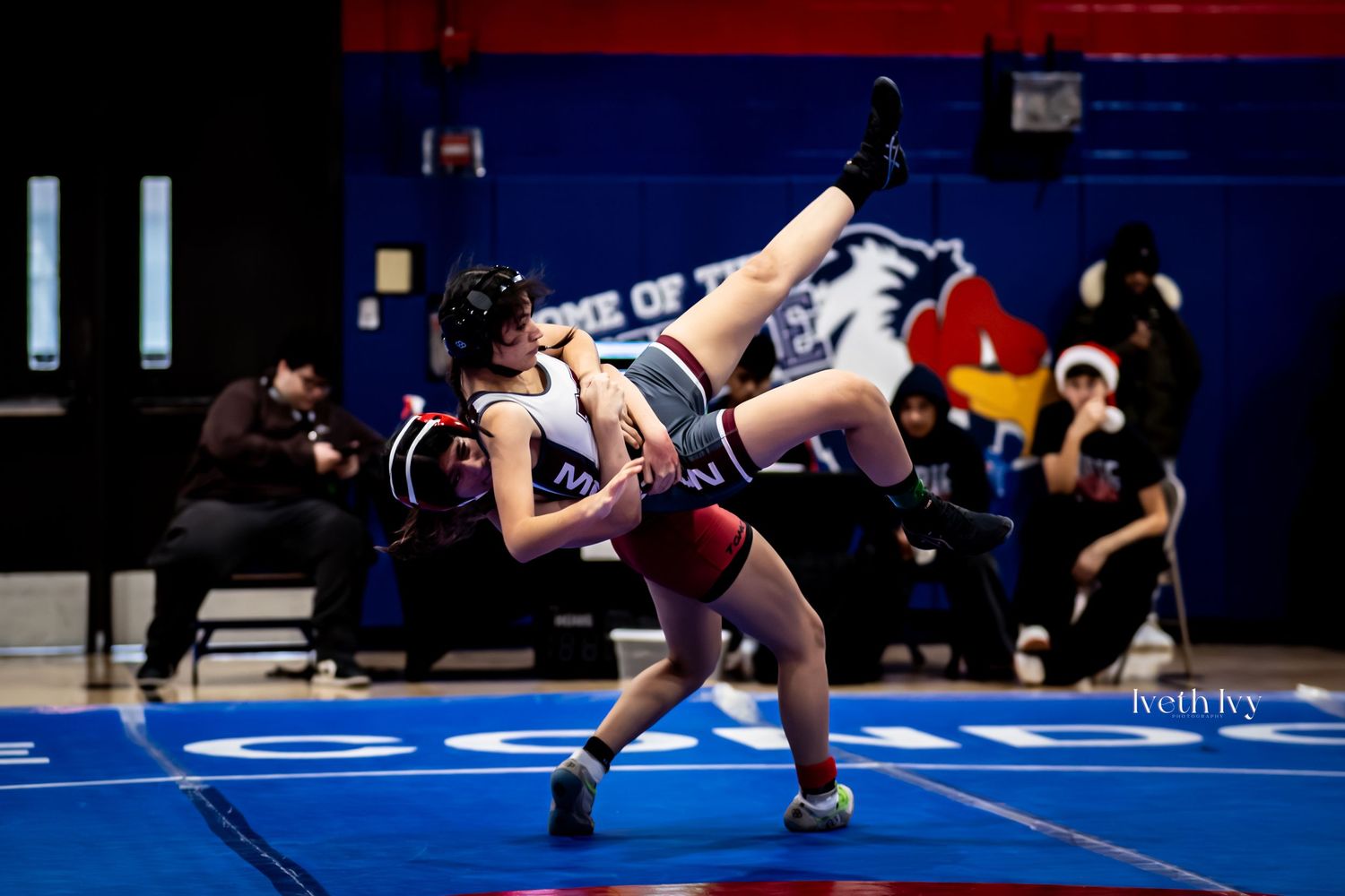 Wrestlers engage in a competitive match on a blue wrestling mat with one attempting a takedown move.