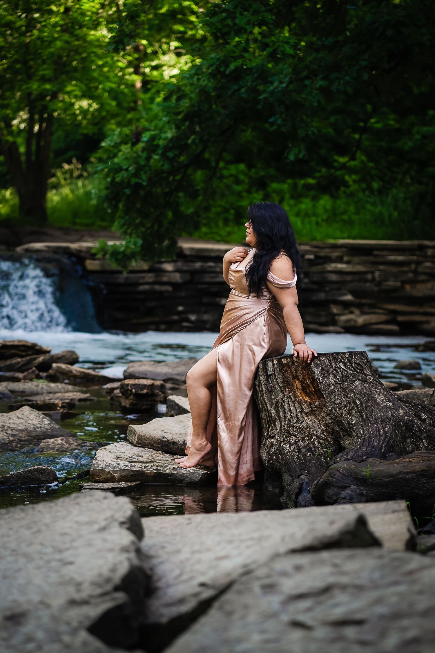 A person sits on rocky outcrops near a cascading river in a serene natural setting surrounded by lush greenery.