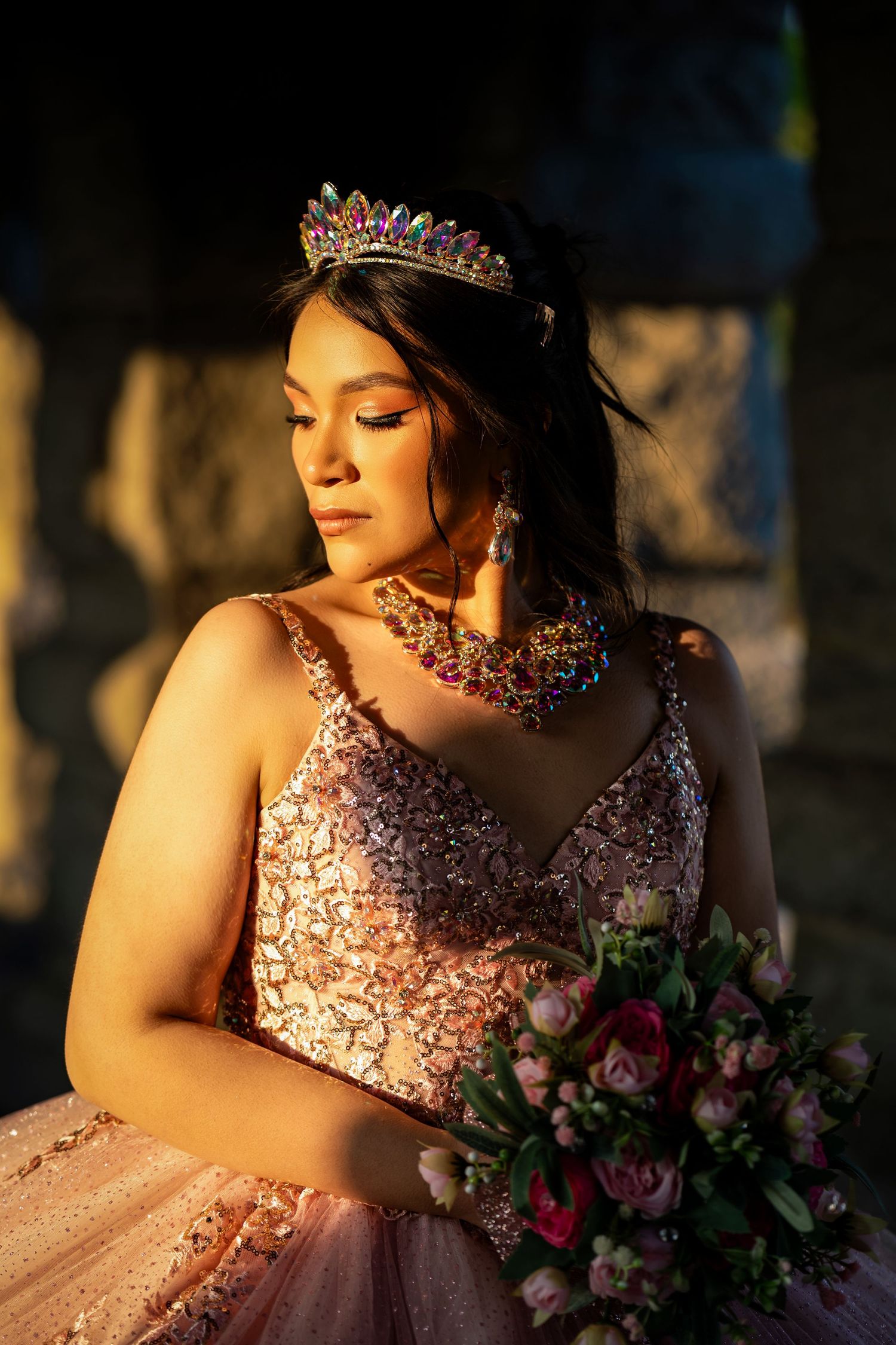 A person in a sparkly pink quinceañera dress poses at golden hour while holding a floral bouquet.