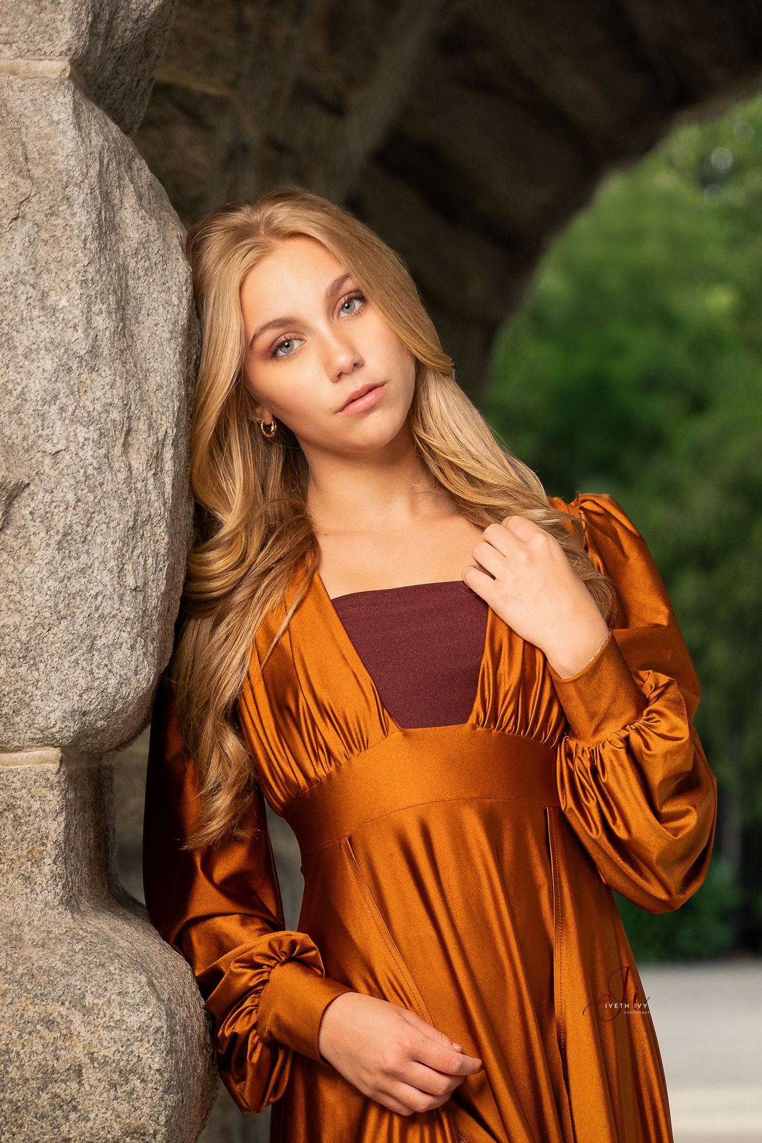 A fashion portrait showcasing a flowing burnt orange dress with long sleeves against a stone column backdrop.