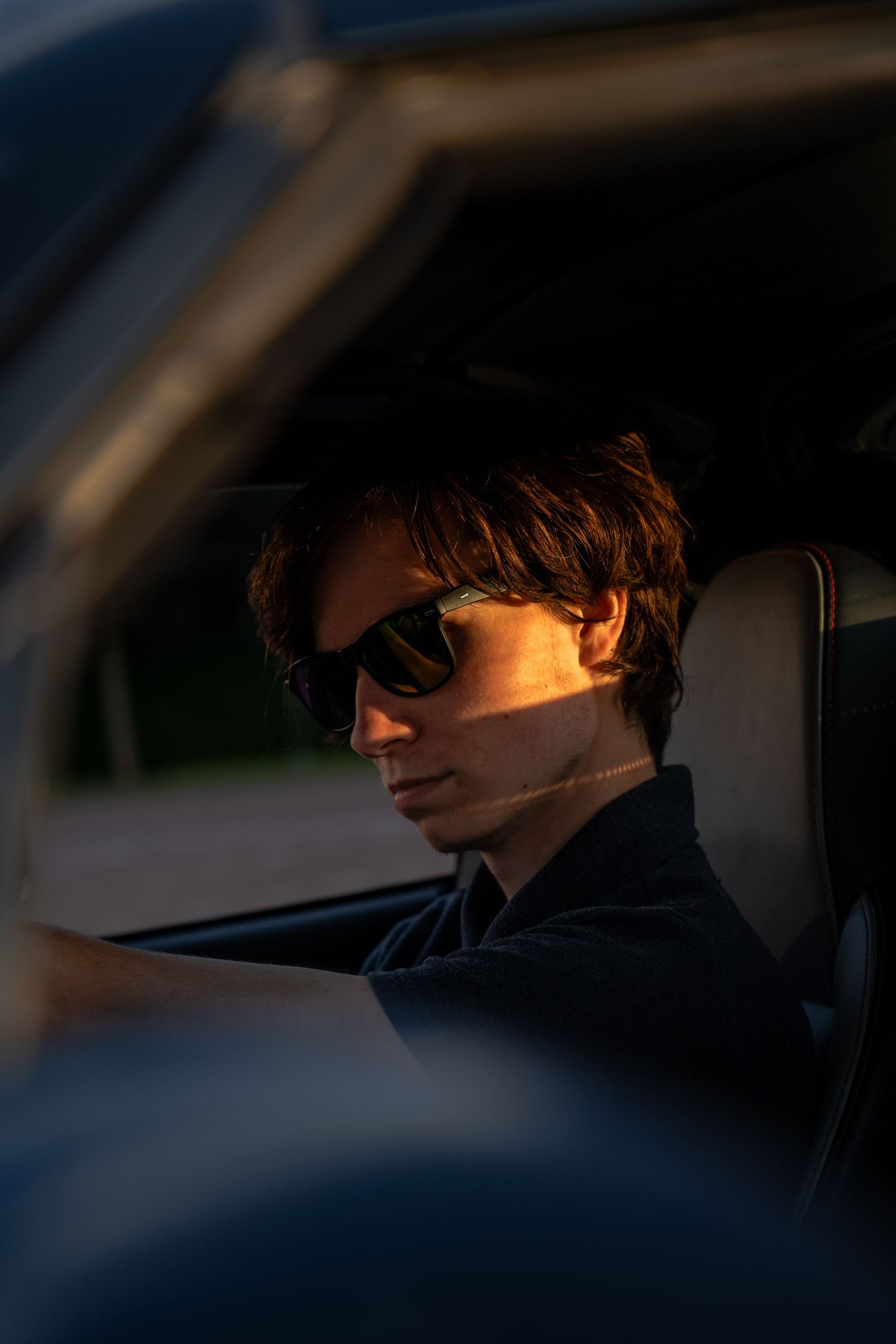 Young man with sunglasses sitting in a car, bathed in warm golden sunset light through the window.