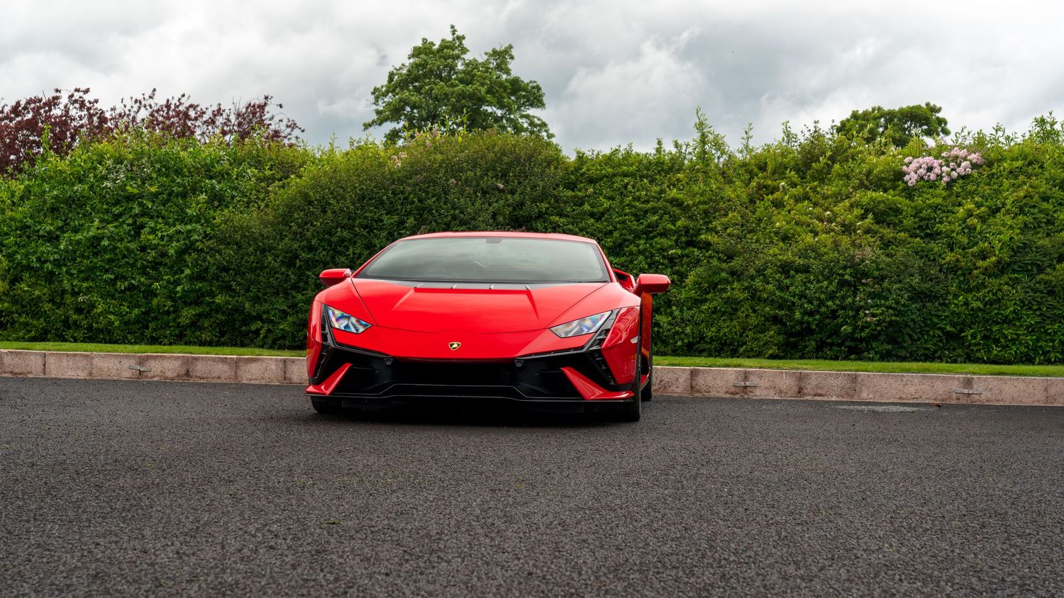 Red Lamborghini Huracan supercar parked on asphalt driveway with lush green hedges in background.