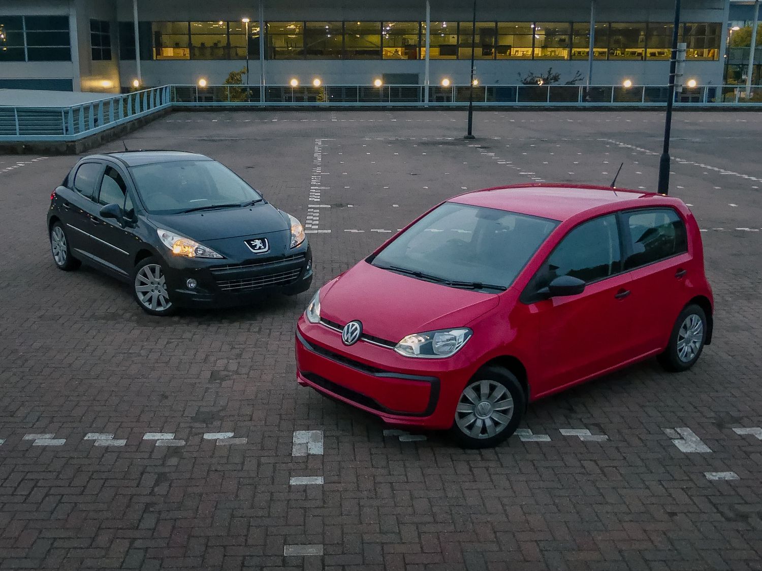 A red Volkswagen Up and a dark blue Peugeot 207 parked together in an empty parking lot at dusk.