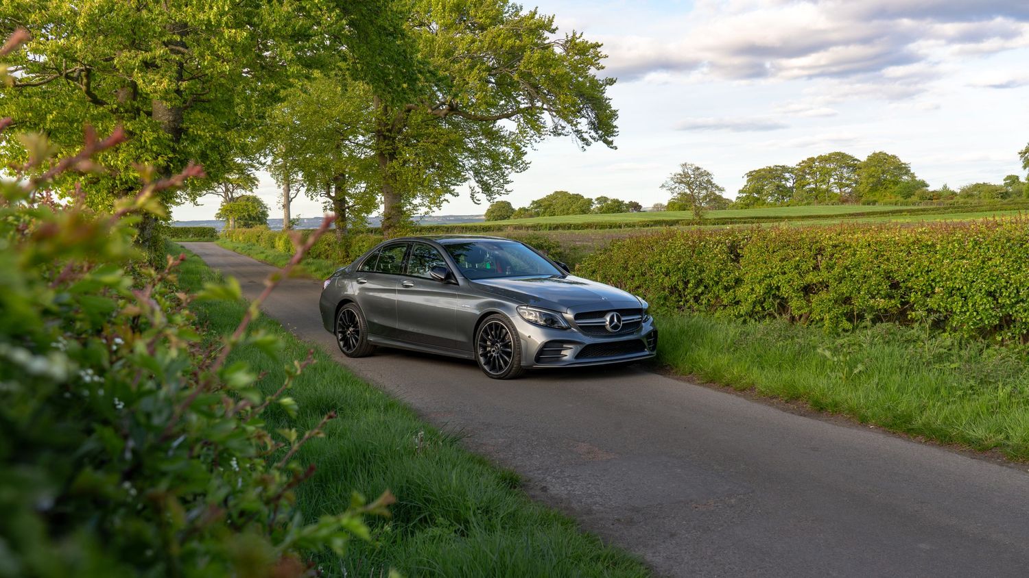 Gray Mercedes-Benz C-Class sedan parked on a narrow country road surrounded by lush green trees and hedgerows.