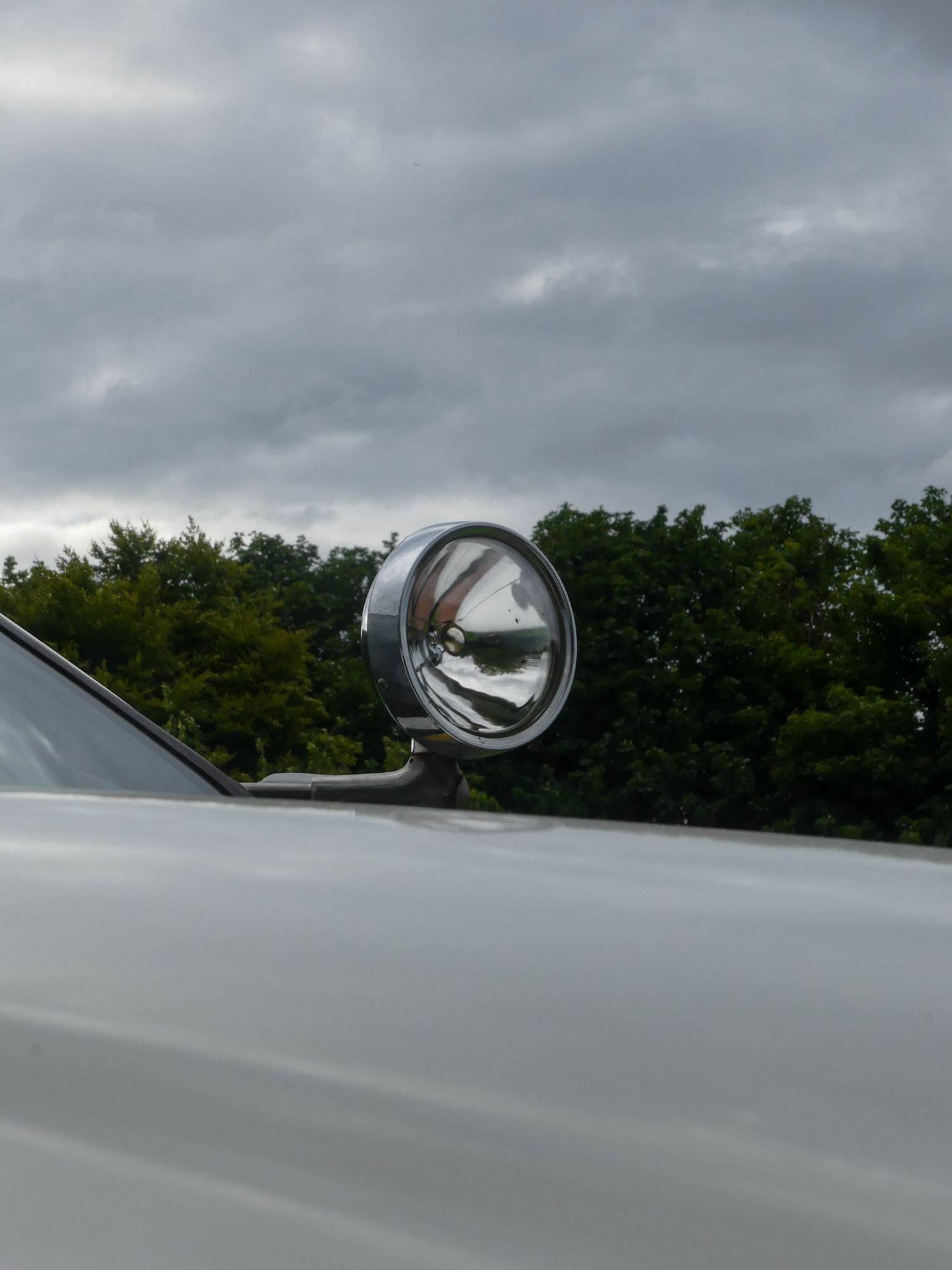 Chrome spotlight mounted on a white vehicle hood against a cloudy sky with trees in the background.