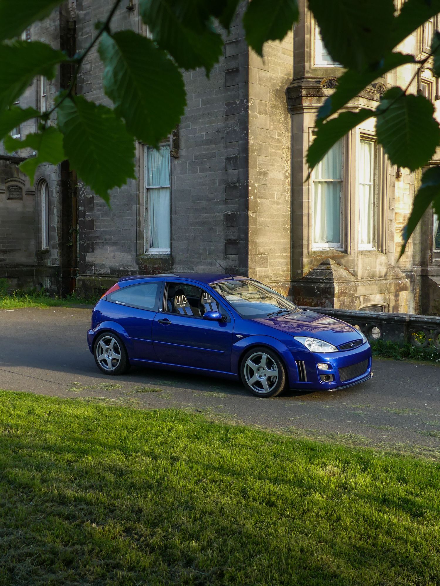 A blue modified Mercedes SL sports car parked on a driveway next to stone buildings in evening light.