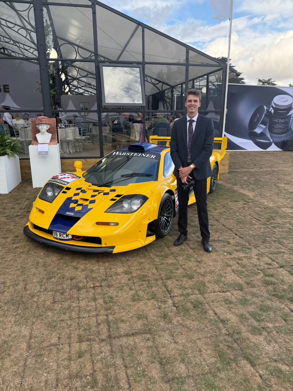 Man in suit posing beside a yellow McLaren F1 GTR Warsteiner racing car at an outdoor motorsport event.