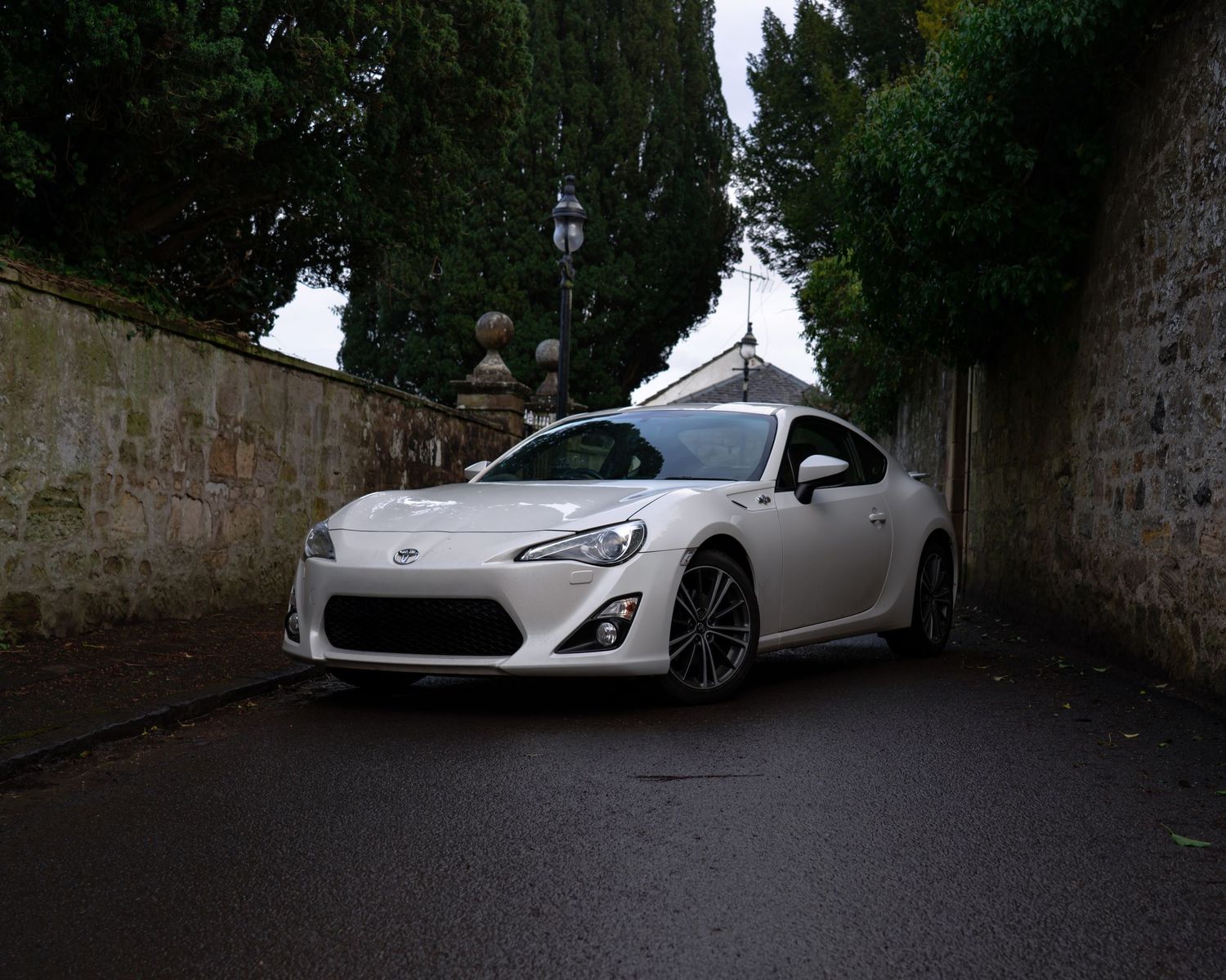 White Toyota 86 sports coupe parked in a narrow stone alleyway surrounded by tall trees and ivy-covered walls.