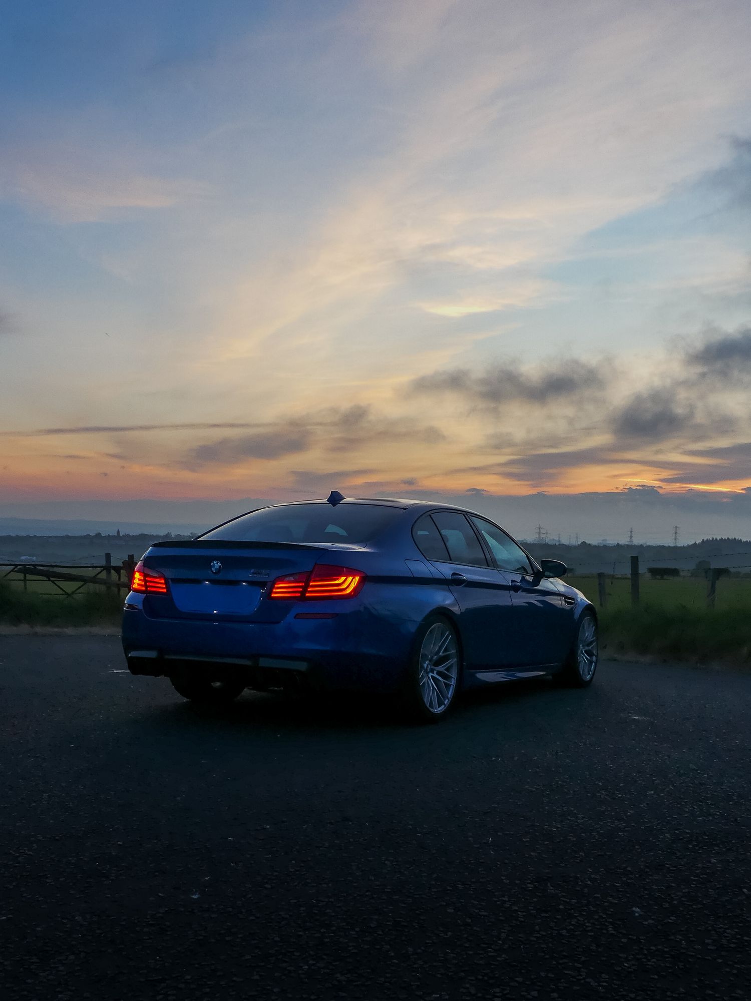 BMW M5 sedan in metallic blue parked on a road during a dramatic sunset with orange and purple sky.