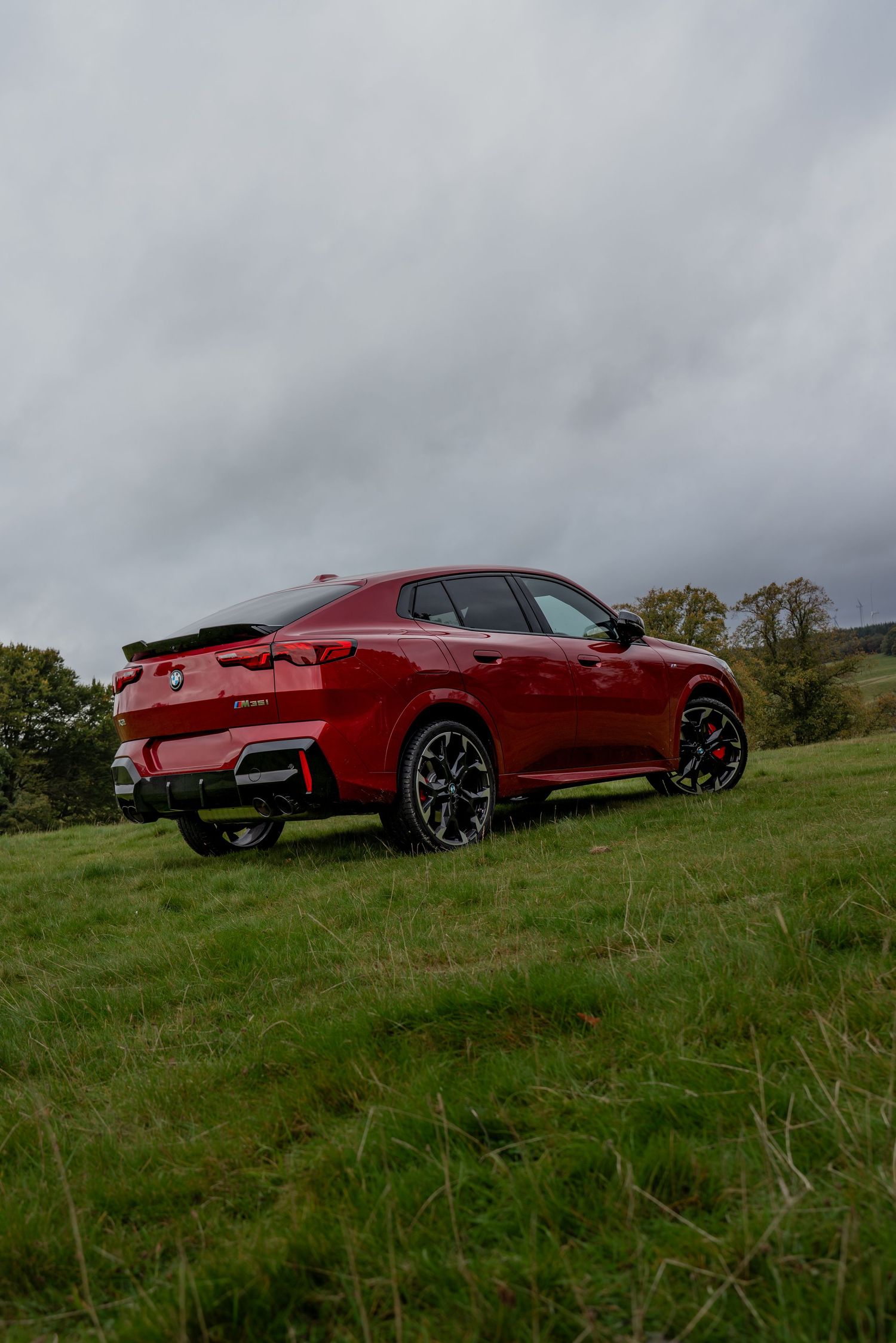 Red BMW X4 M Competition SUV parked on grassy hill against cloudy sky showing sporty profile and distinctive wheels.