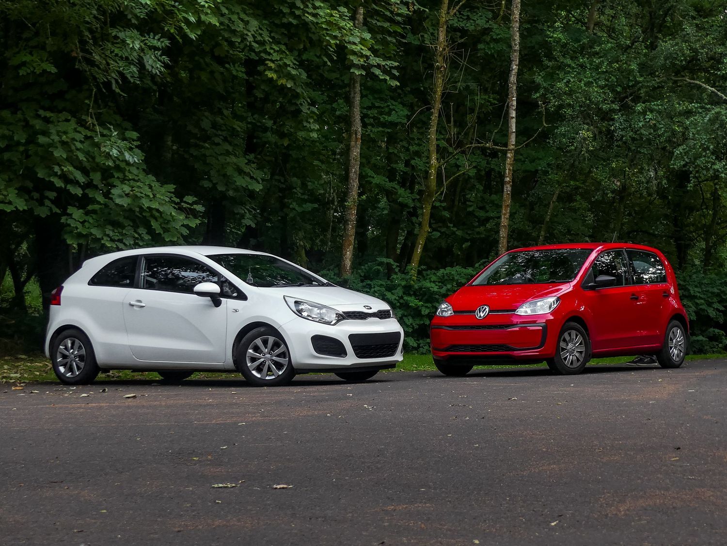Red VW UP! and white Kia Rio hatchback cars parked on a forest road with green trees in the background.