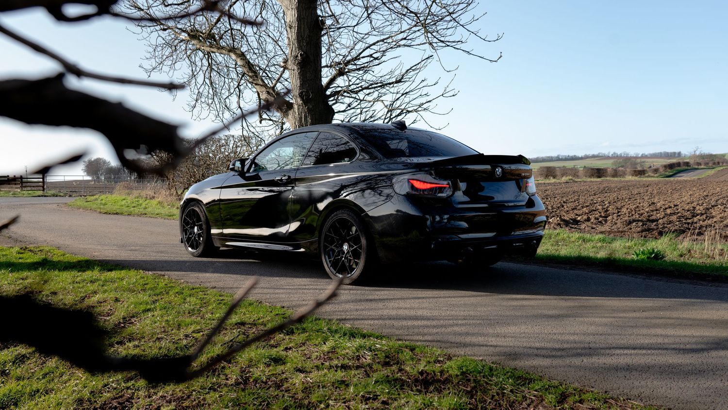 Black BMW M2 coupe parked on a rural road surrounded by green fields and bare trees on a clear day.