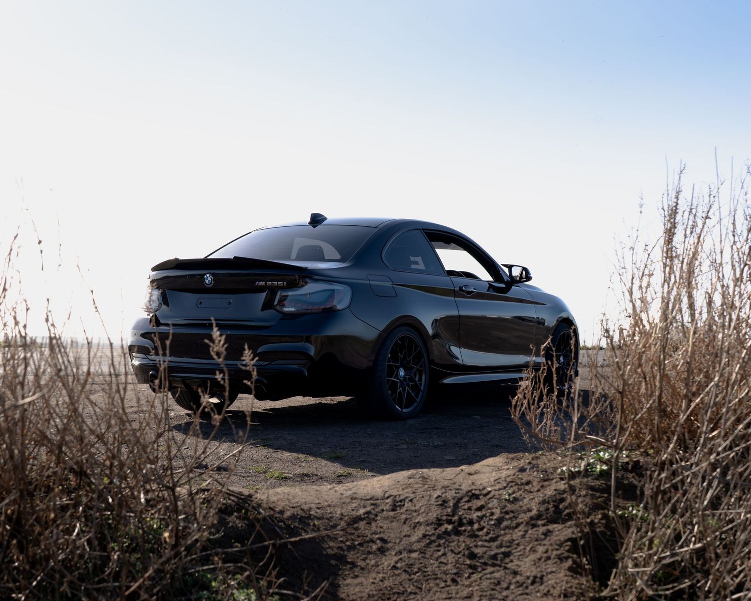 A sleek black BMW M2 coupe parked on a dirt trail surrounded by dry grass under a clear sky.