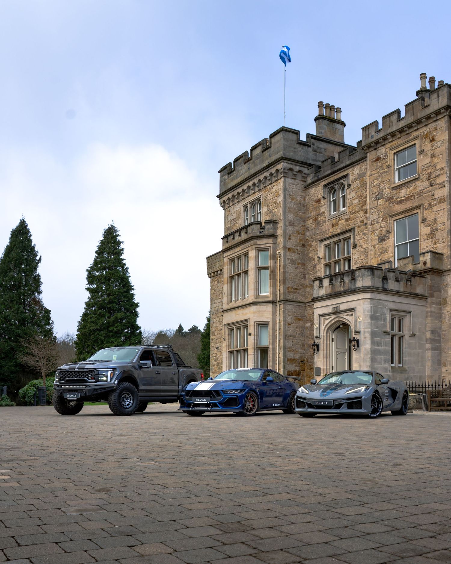 Three luxury vehicles parked in front of a grand stone castle with Gothic architecture and towering evergreen trees.