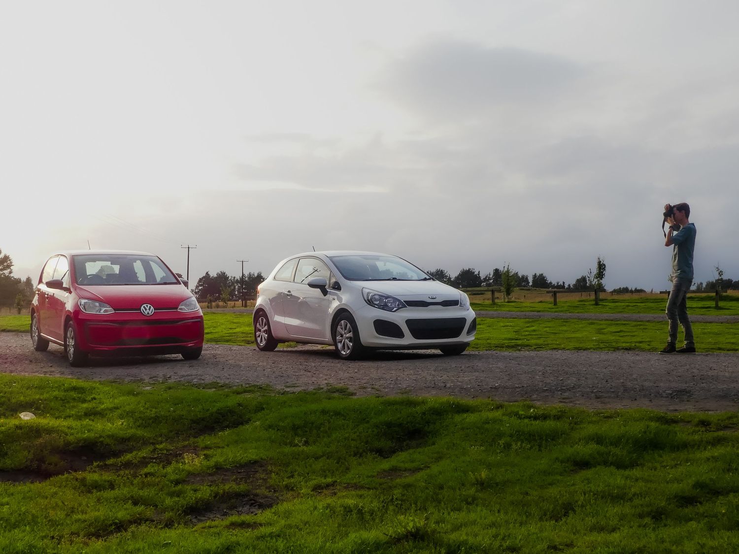 Two cars parked on grassy hilltop with a photographer taking pictures against cloudy sky backdrop.