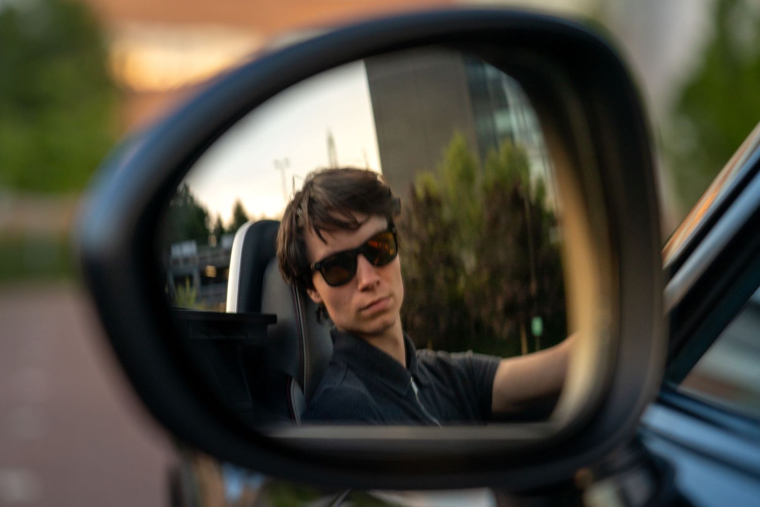 Person with sunglasses reflected in a car side mirror, with trees and buildings visible in the background.