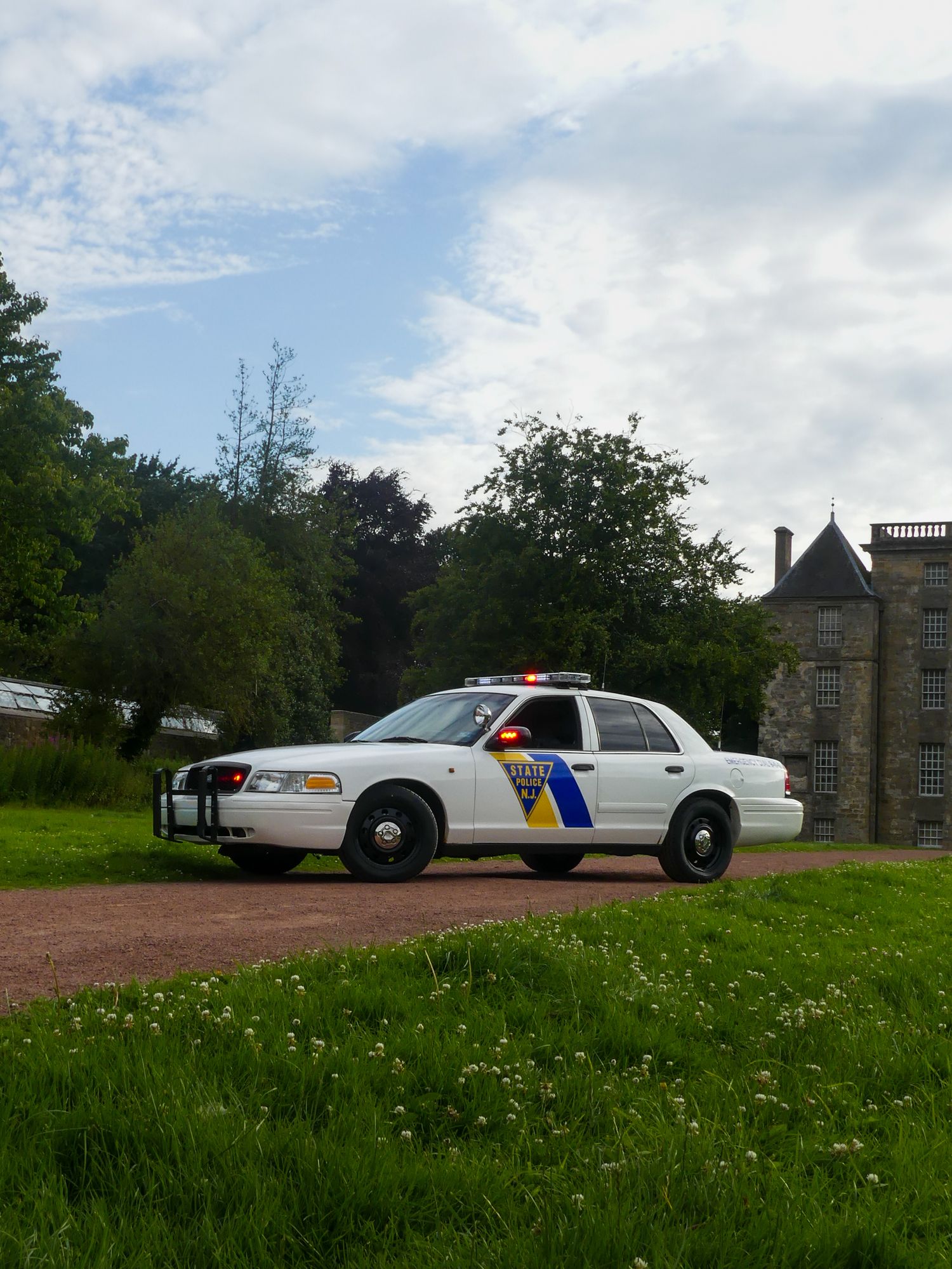 A white police patrol car with emergency lights stationed on a dirt path near a historic stone building.