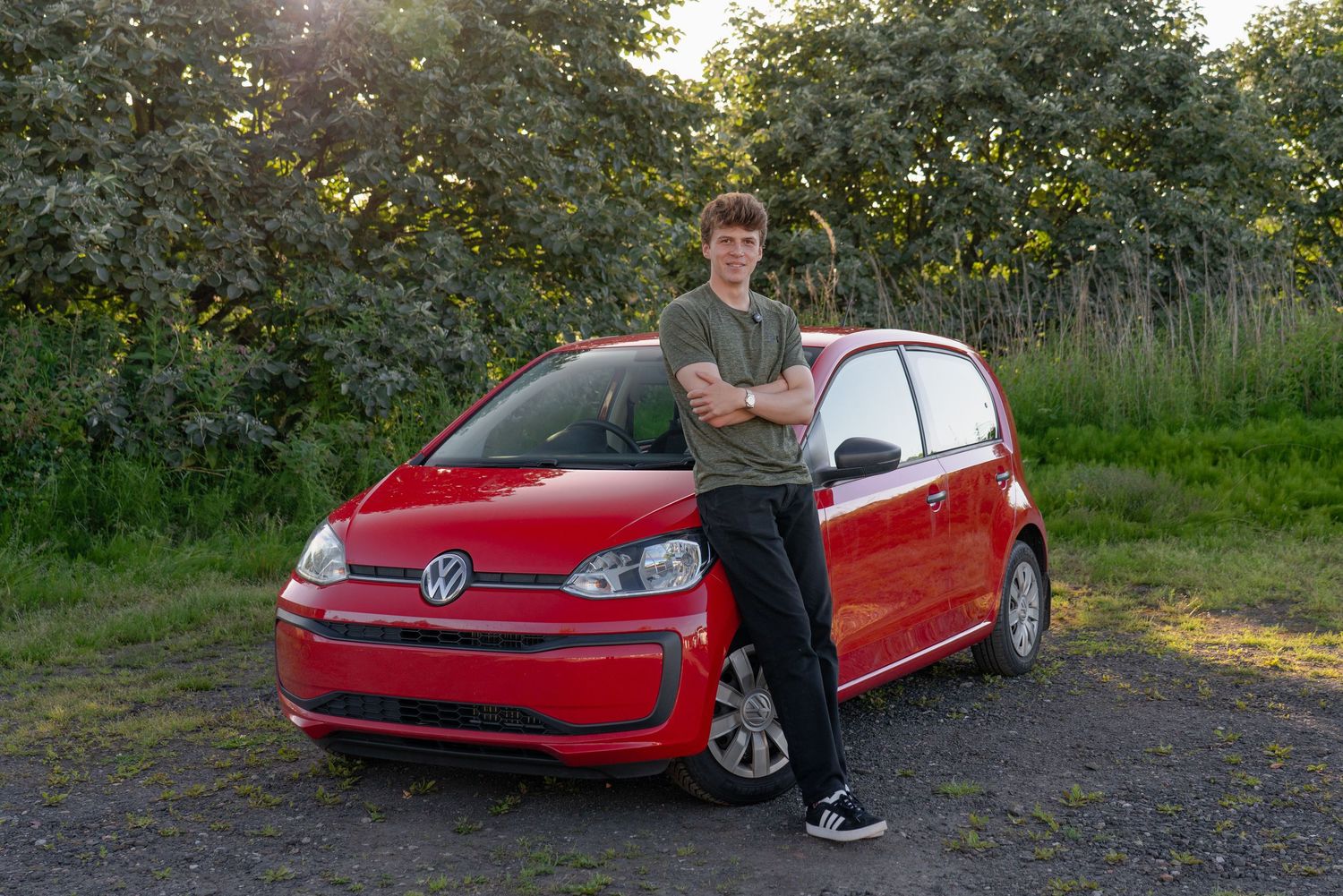Young man leaning against a red Volkswagen Up hatchback parked on a gravel path surrounded by green trees.