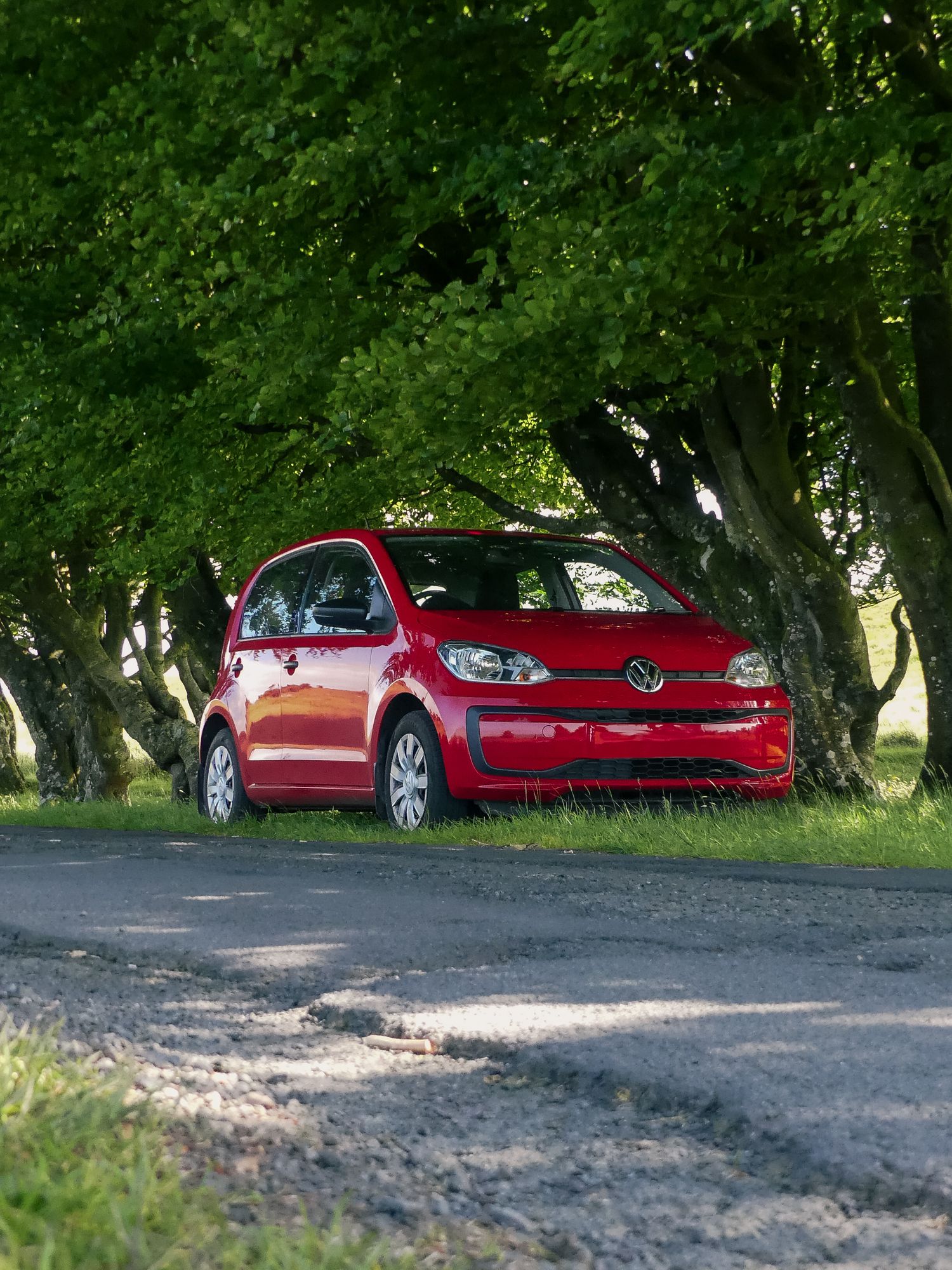 A red Volkswagen Up city car parked on a rural road surrounded by lush green trees in natural lighting.