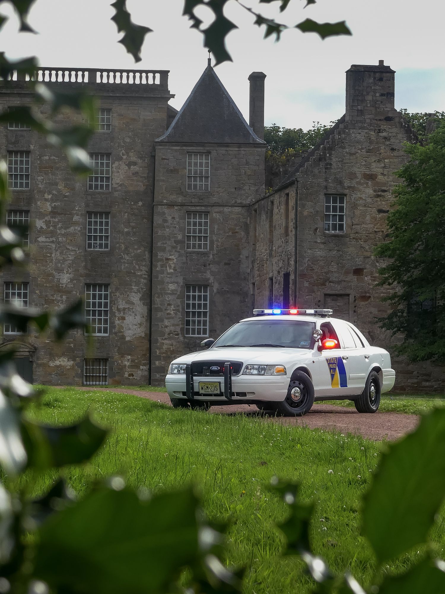 Police car with flashing lights viewed through foliage near historic stone building.