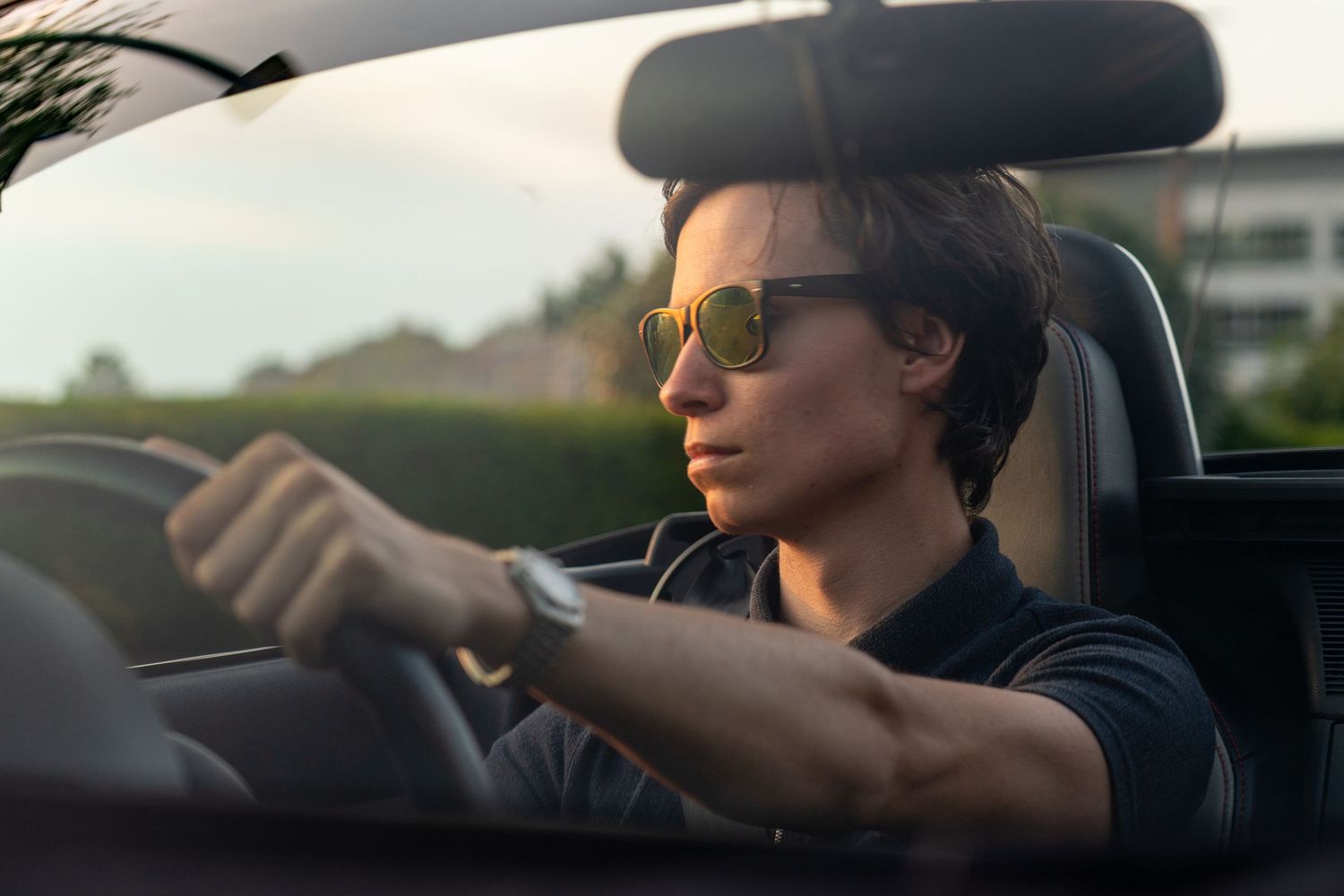 A focused woman wearing sunglasses drives a convertible car, gripping the steering wheel with both hands.