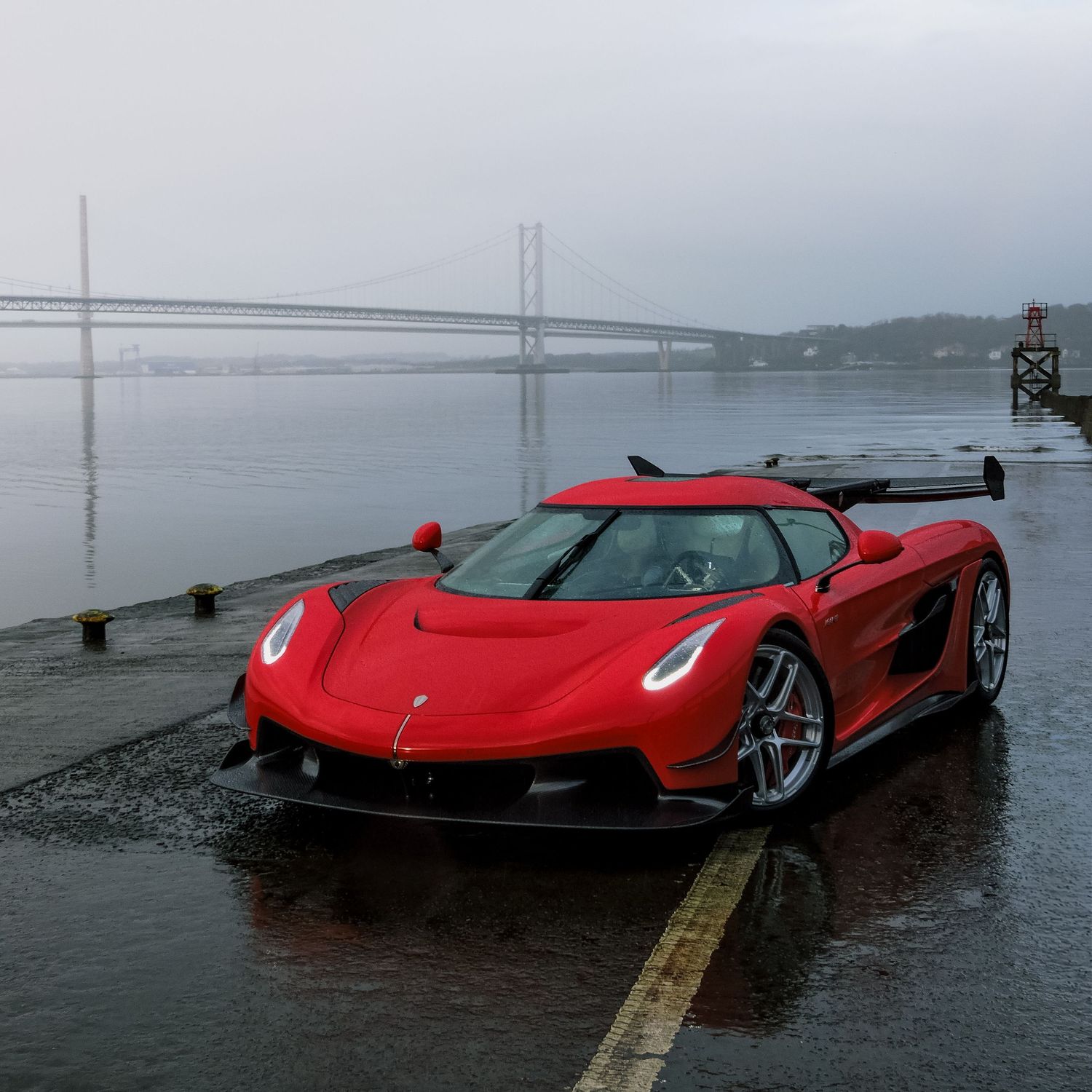 A red Koenigsegg Jesko supercar parked by the waterfront with a bridge visible in the foggy background.