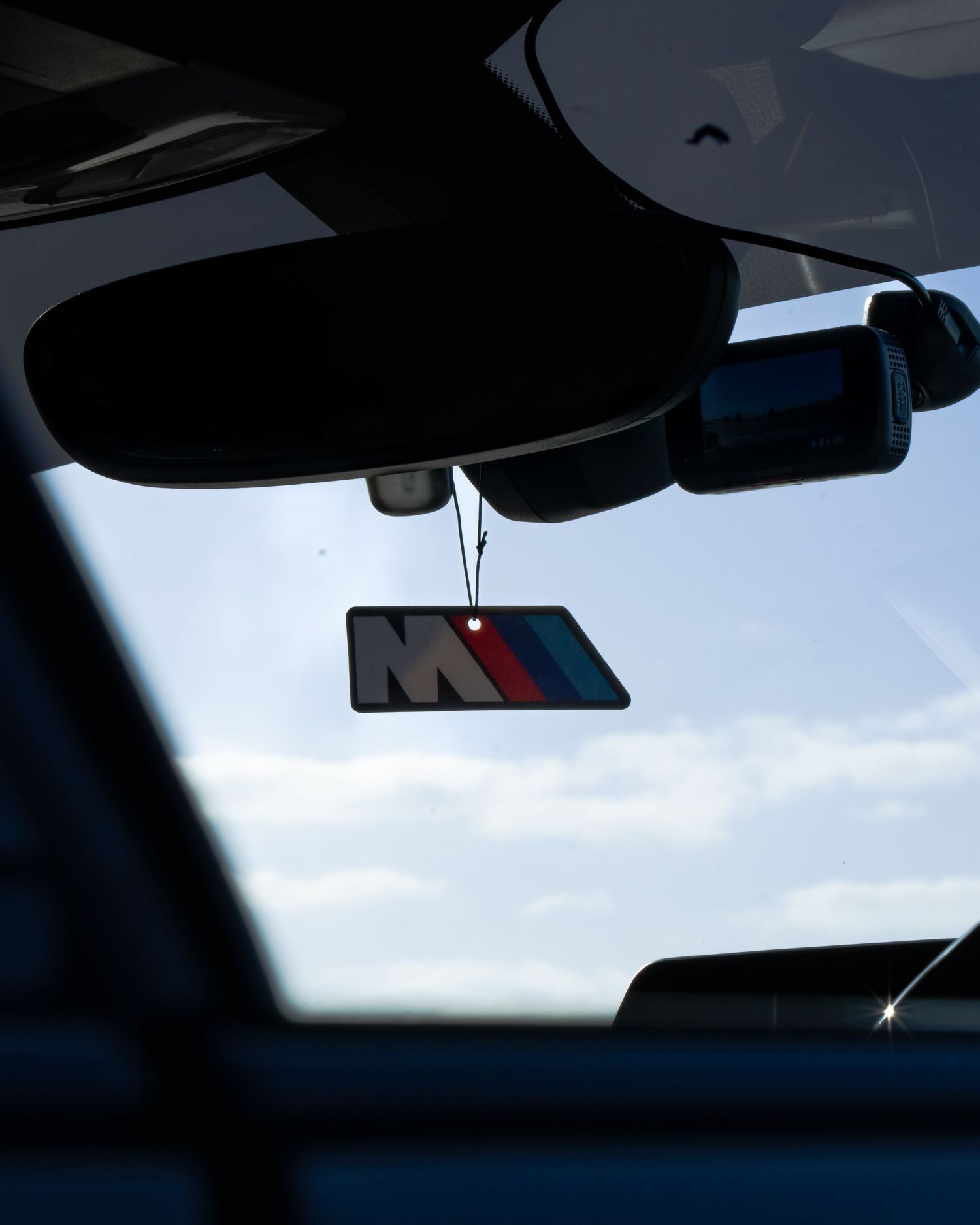 BMW M Series air freshener hanging from rearview mirror inside a car with blue sky visible through windshield.