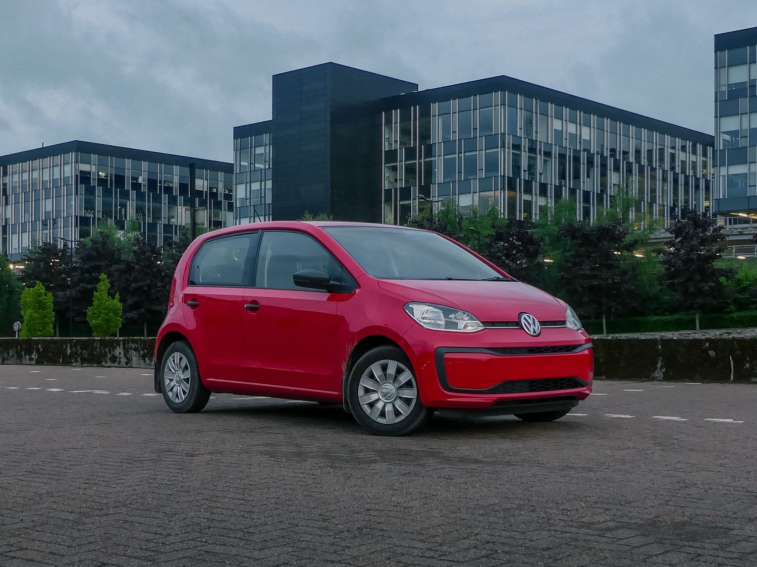 A red Volkswagen up! compact car parked in front of modern office buildings on an overcast day.
