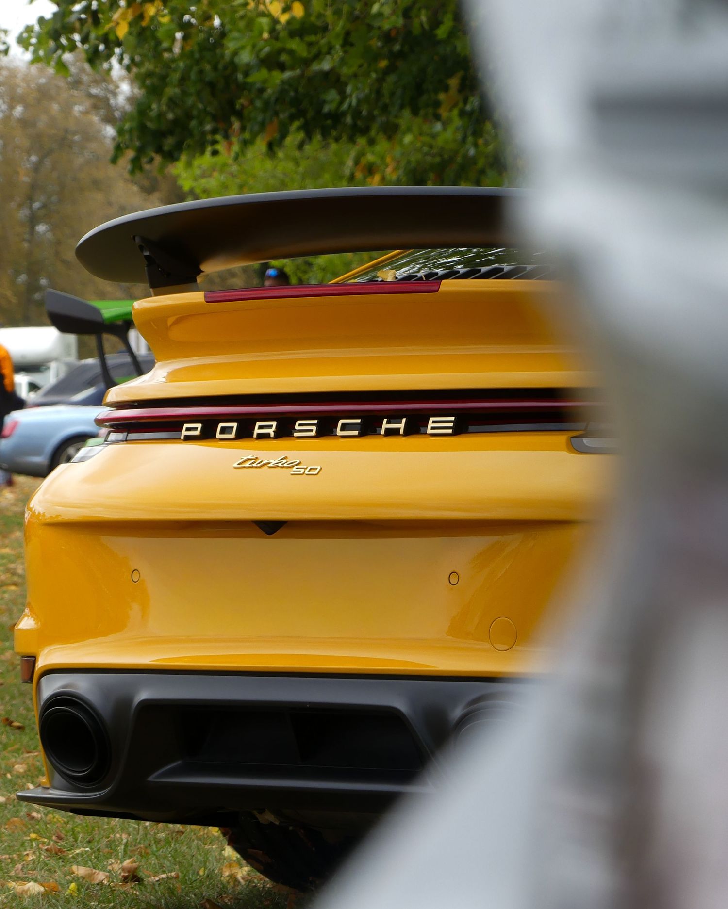 Yellow Porsche sports car rear end detail showing spoiler and exhaust against autumn foliage background.