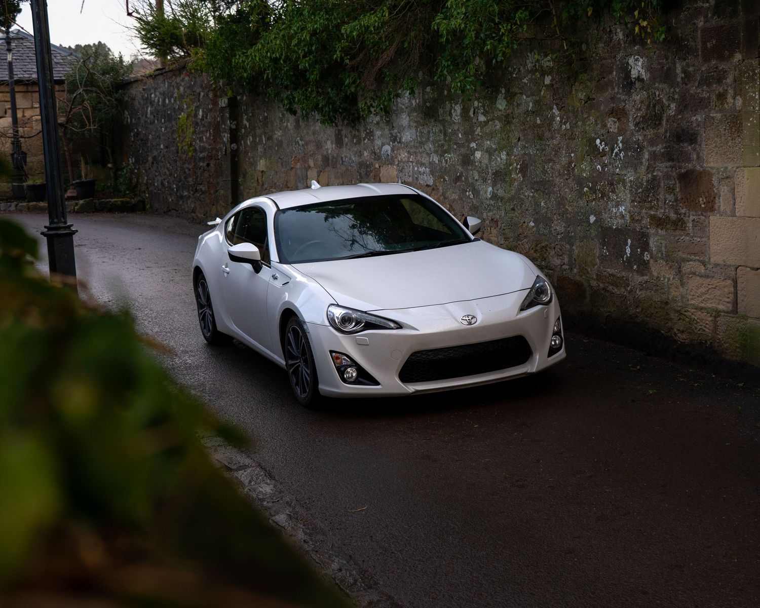 White Toyota 86 sports car parked on a wet narrow road beside a stone wall surrounded by lush green trees.