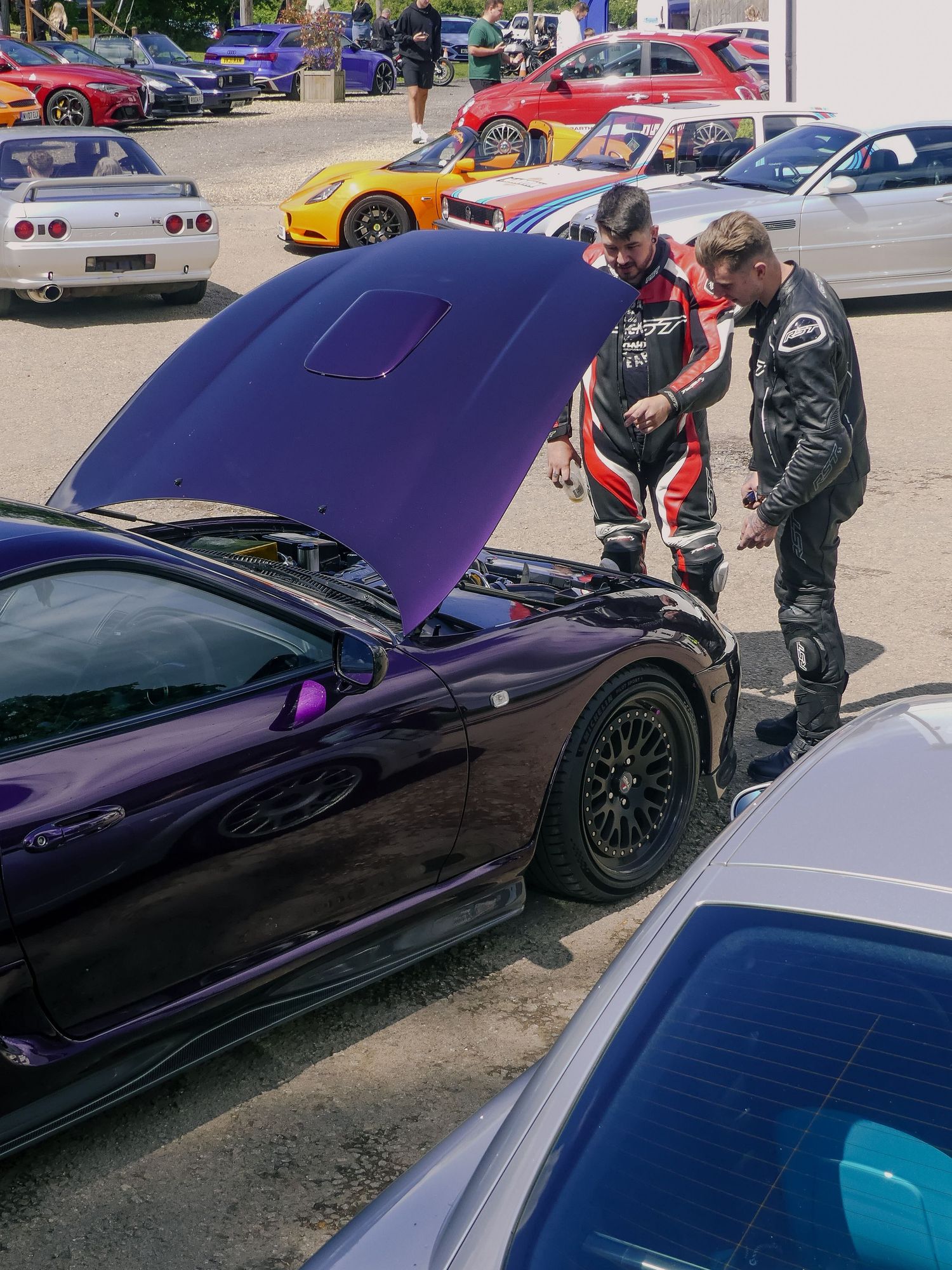 Two car enthusiasts examine under the hood of a purple sports car at an auto show with other vehicles in the background.
