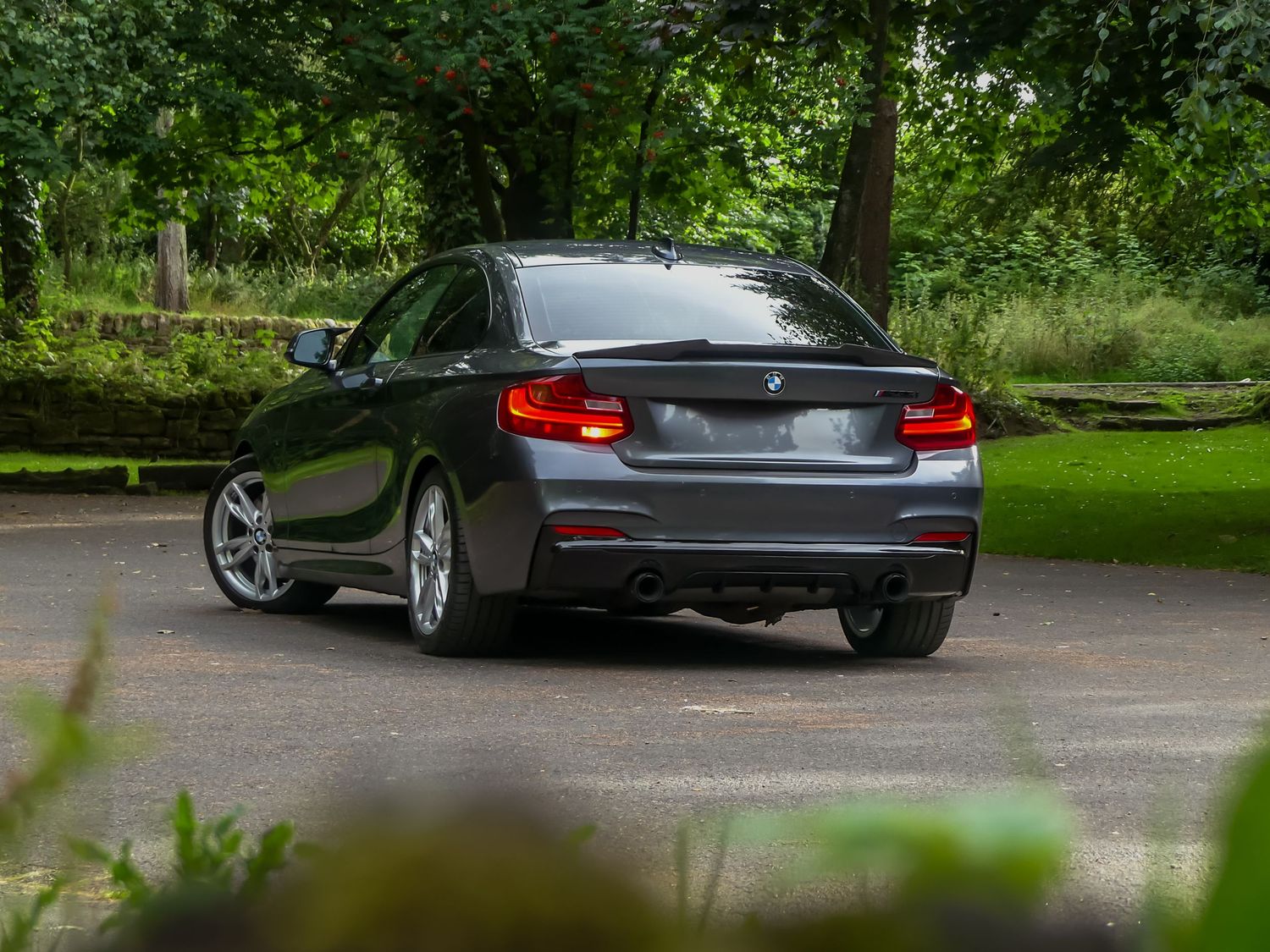 A sleek grey BMW 3-series sedan parked on a tree-lined driveway with green foliage in the background.