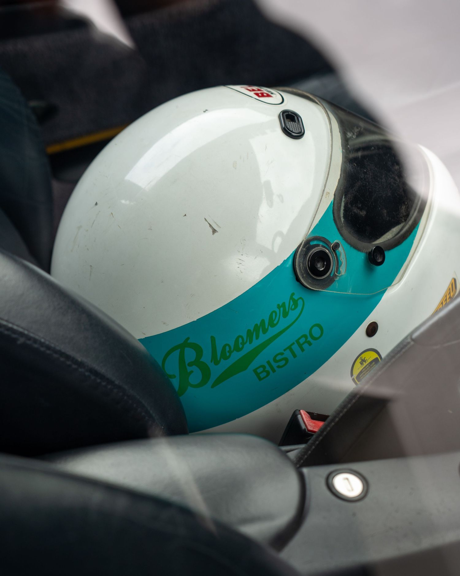 White and teal racing helmet with Bloomers Bistro logo resting in a race car seat.