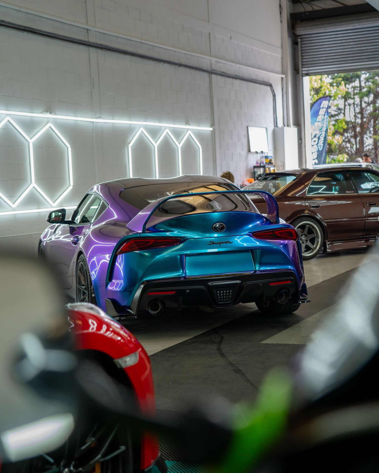 A purple and teal Toyota Supra GR parked inside a modern car garage with geometric neon lights on the wall.