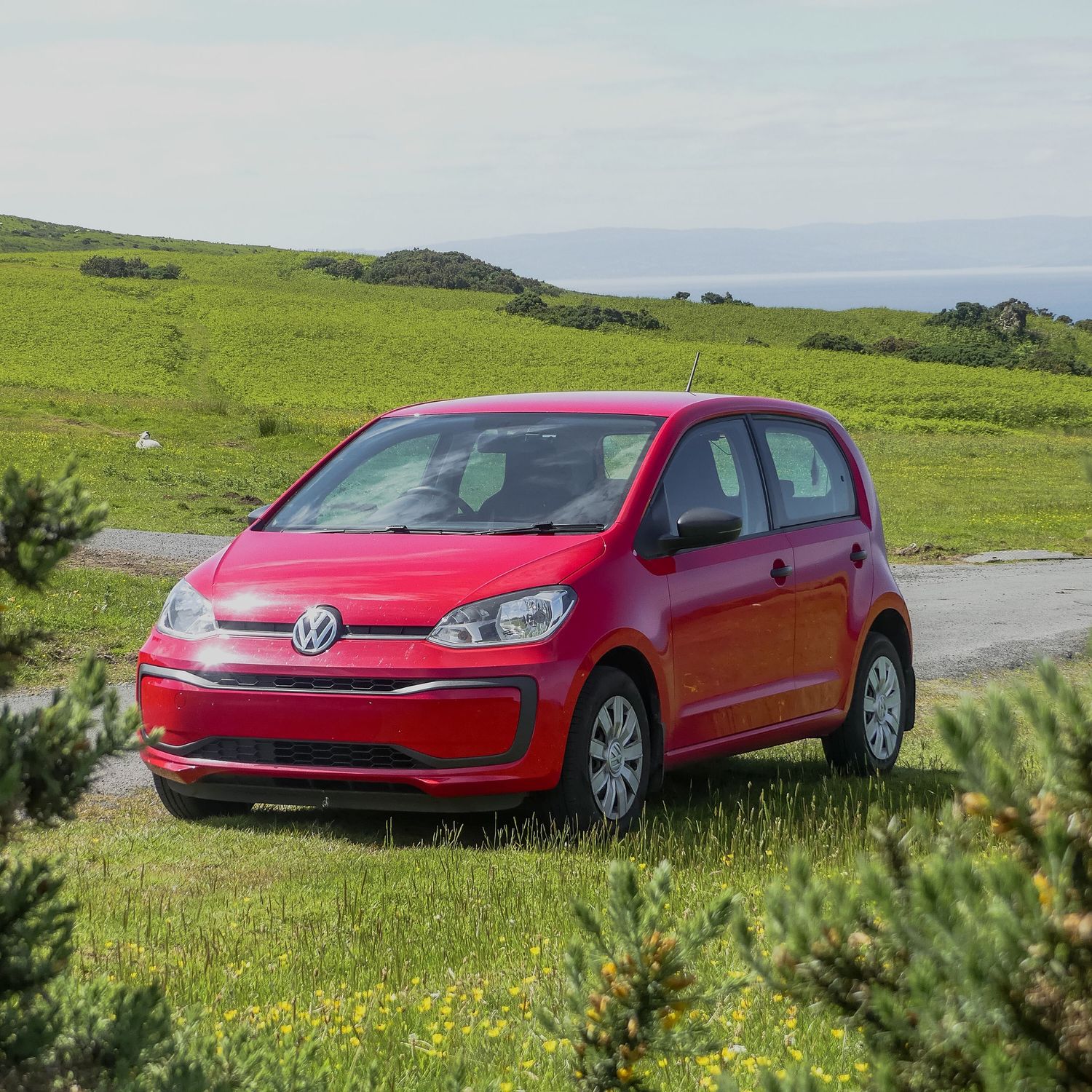 A red Volkswagen Up compact car parked on a scenic coastal hill with green grass and ocean views in the background.
