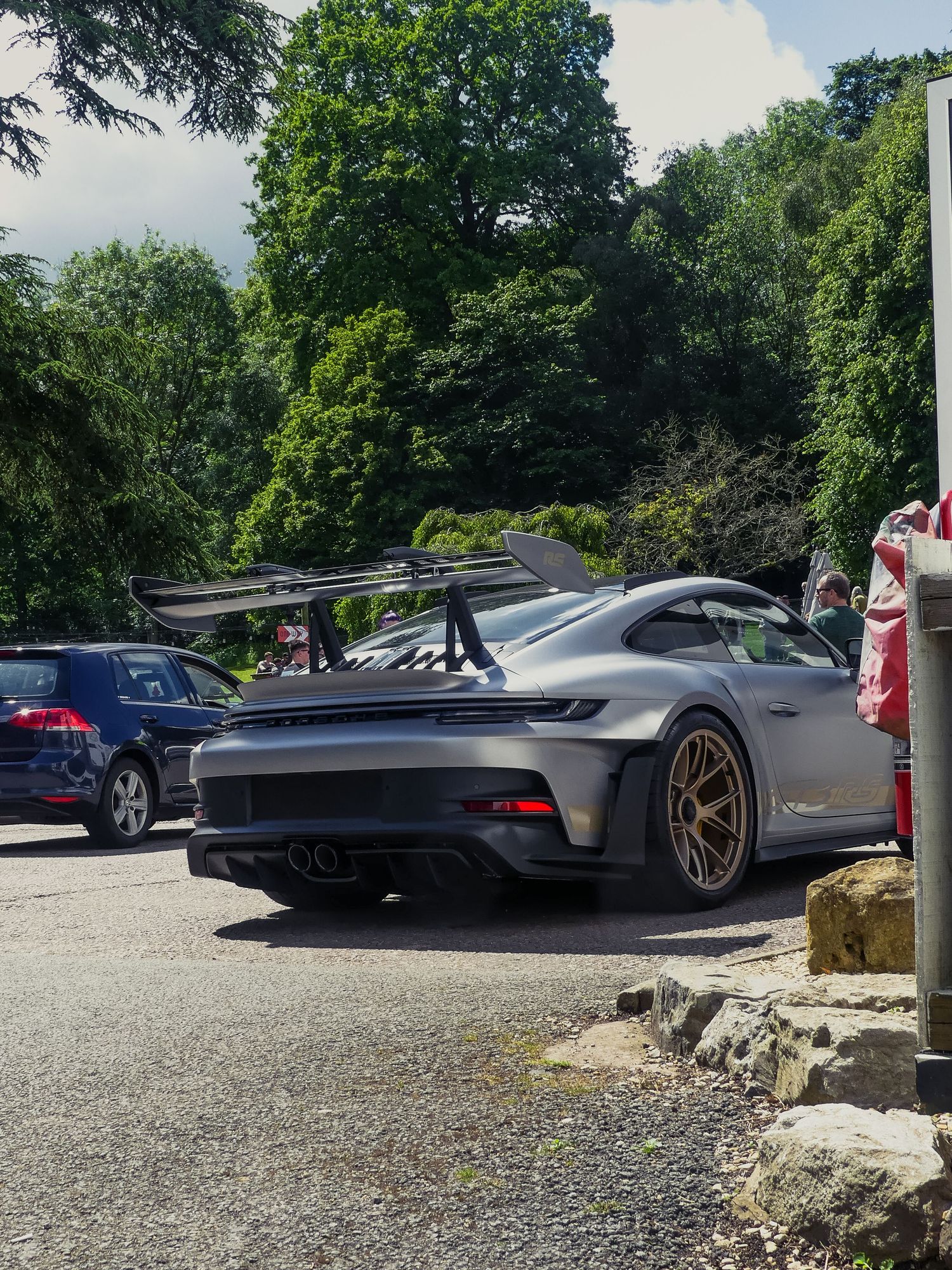 A silver Porsche supercar parked in a gravel lot with other vehicles and trees visible in the background.