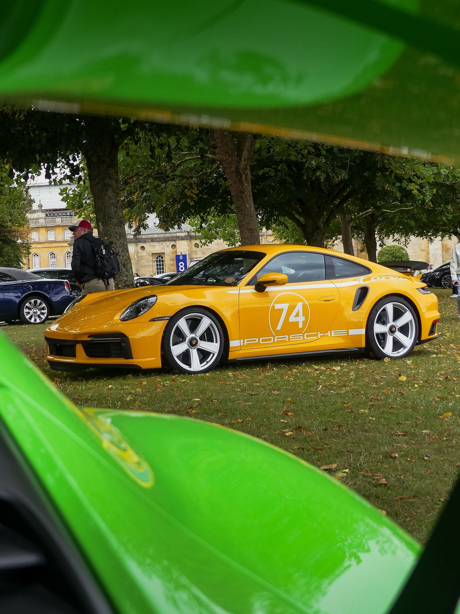 Yellow sports car with racing number 74 and custom wheels parked on grass under trees.