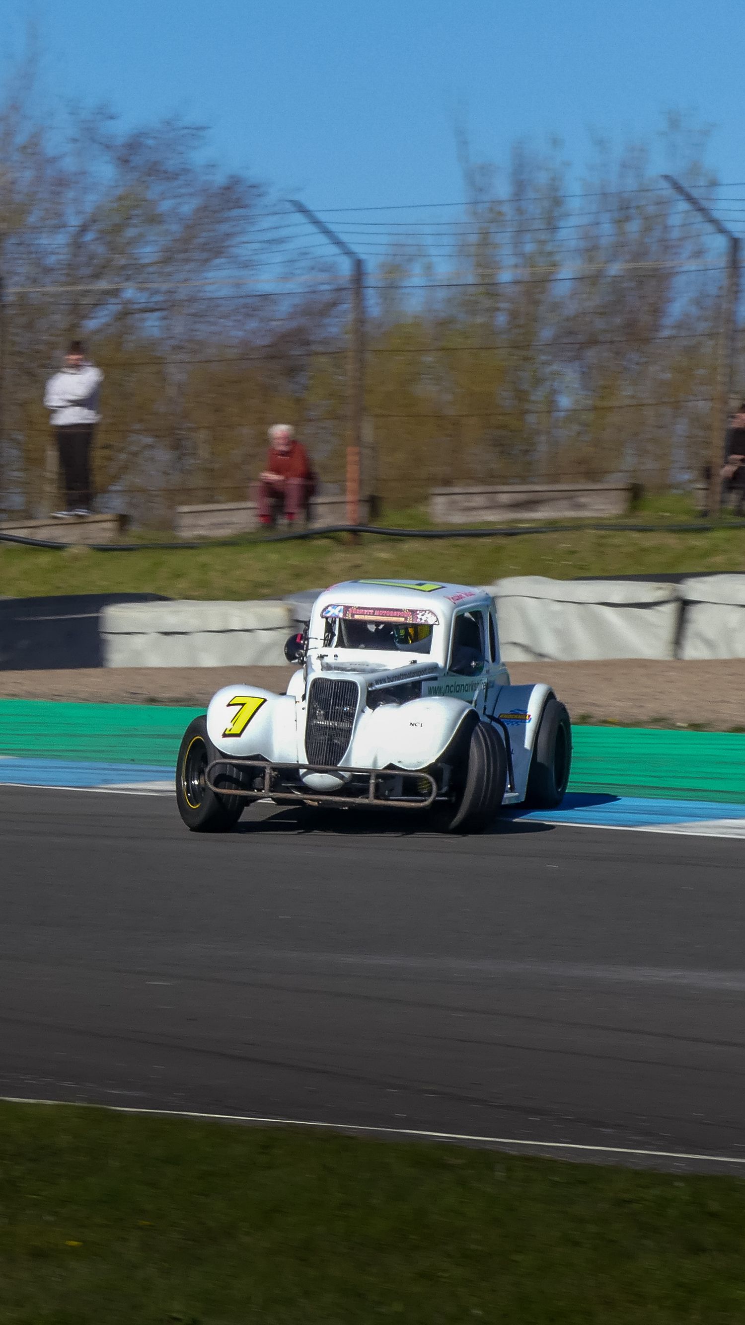 A white vintage racing car with number 7 speeds around a racetrack on a sunny day.