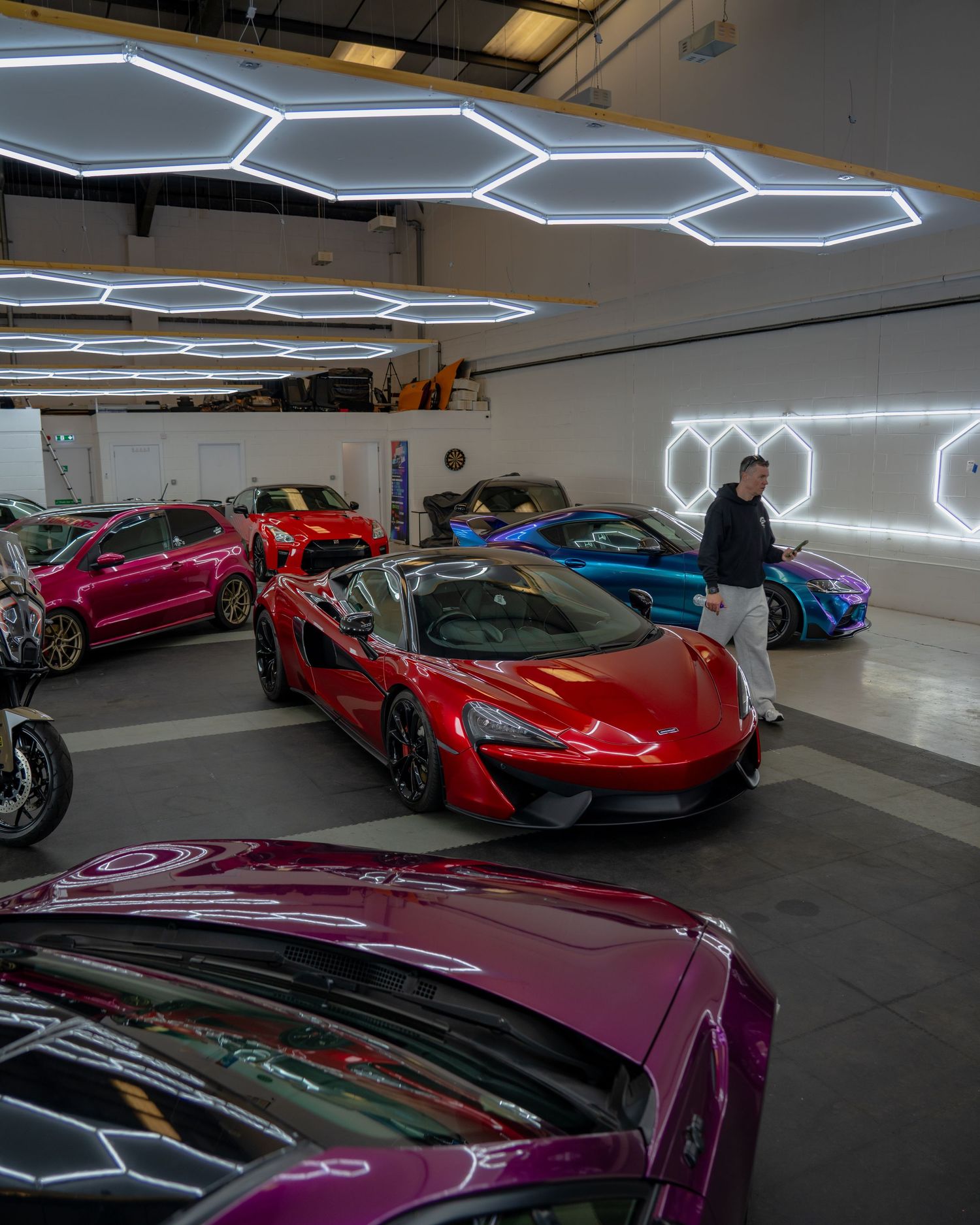 Luxury supercar showroom featuring McLaren and exotic cars under hexagonal LED lights with a man browsing the collection.