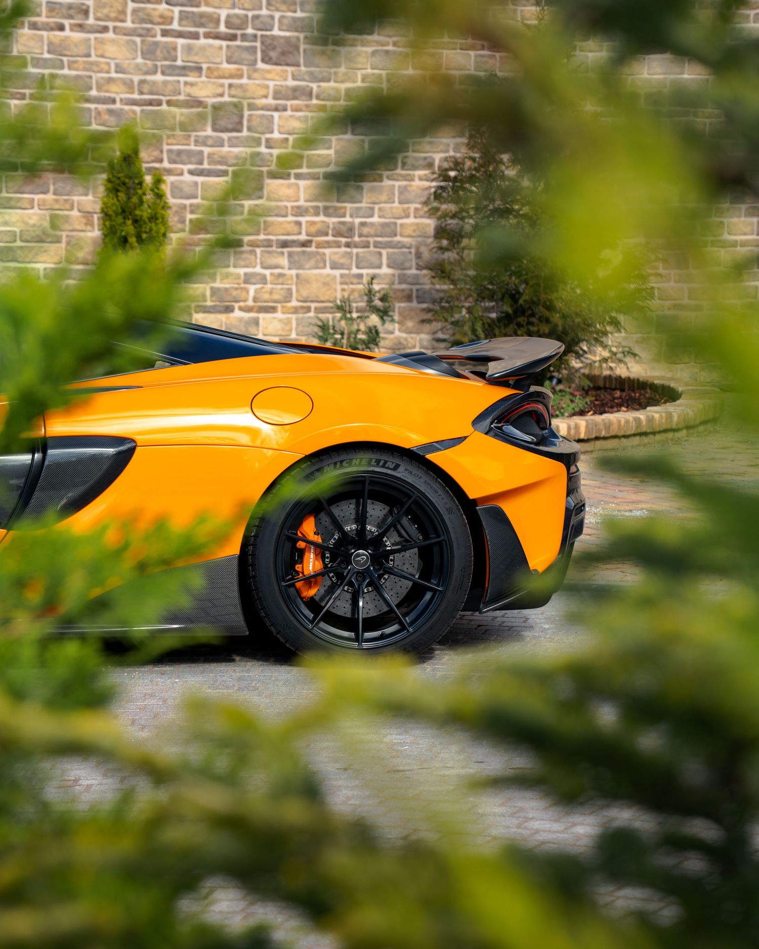 Orange McLaren supercar rear view with active spoiler, seen through lush green foliage against a stone wall backdrop.