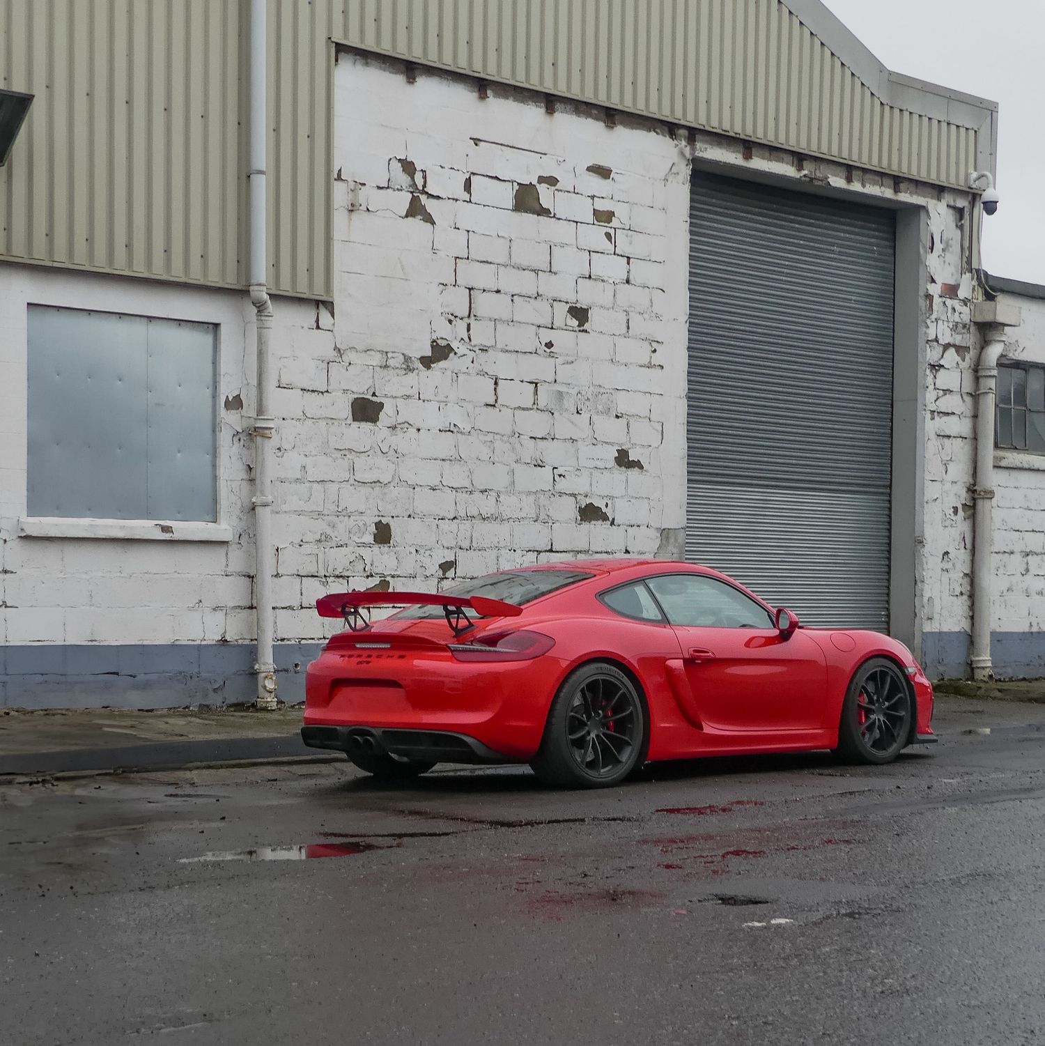 Red sports car parked outside industrial warehouse buildings in wet conditions.
