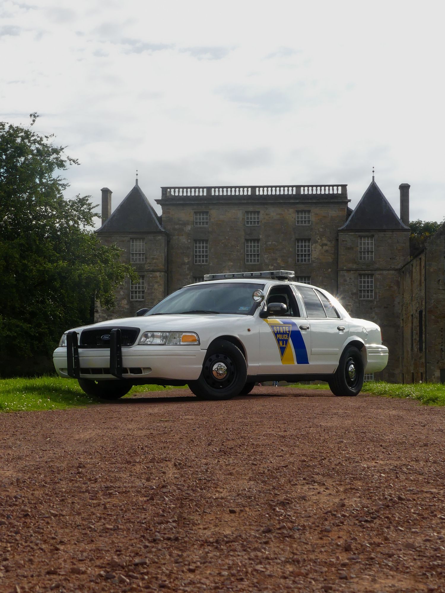 A Ford Crown Victoria police car parked on a gravel path leading to a historic stone mansion.