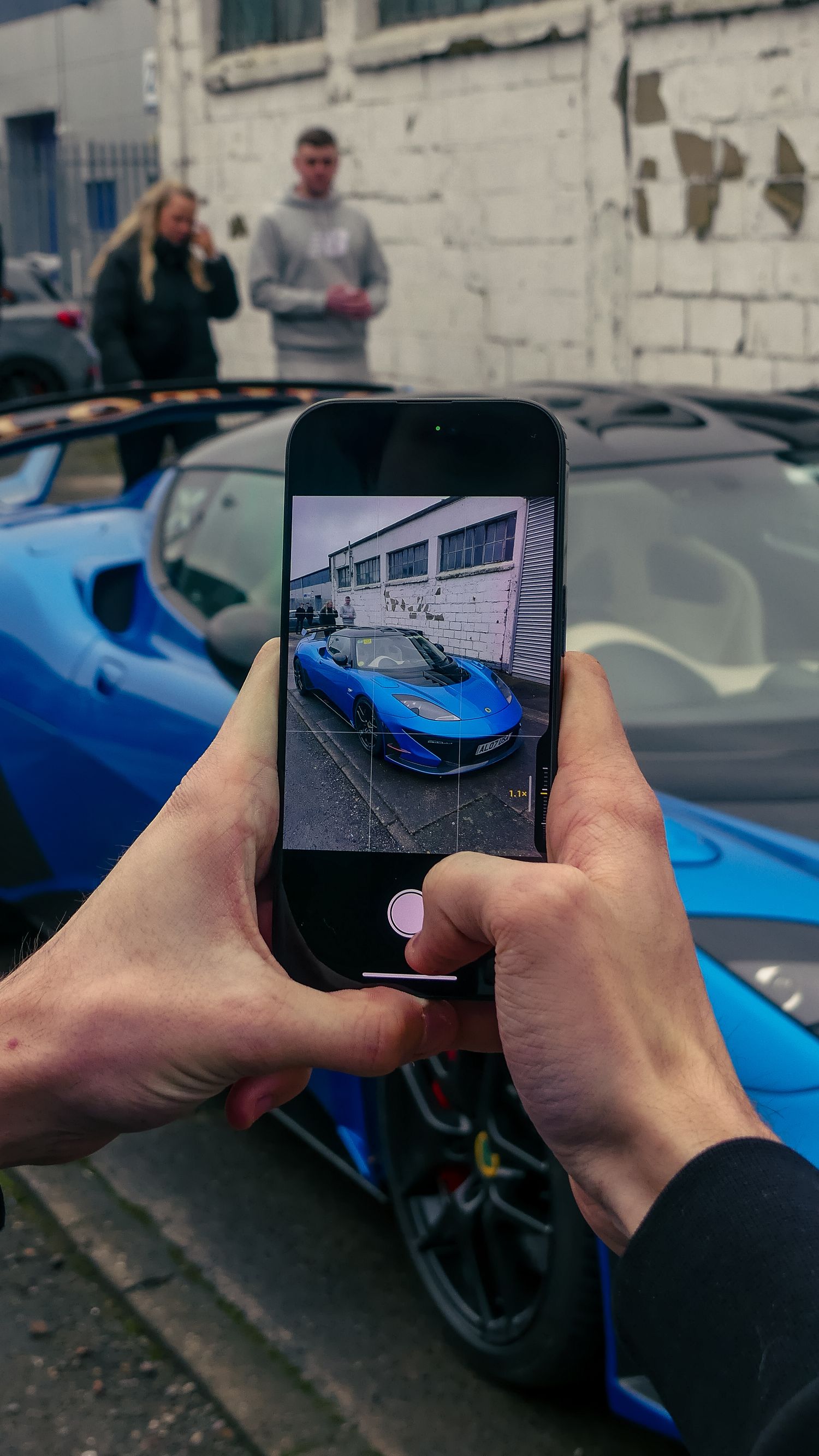Someone takes a photo of a blue sports car on their smartphone while other people stand in the background.
