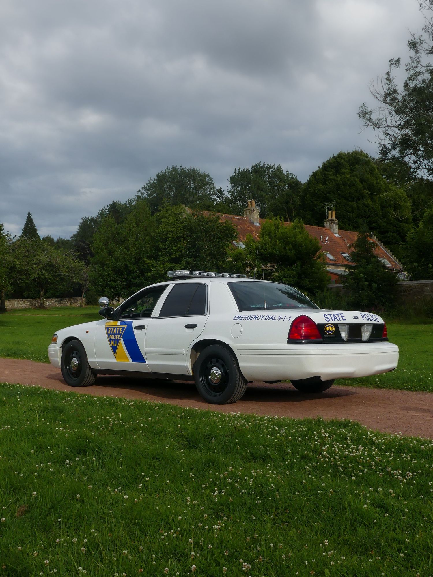 New Jersey State Police Crown Victoria parked on a dirt path with red-roofed buildings visible in background.