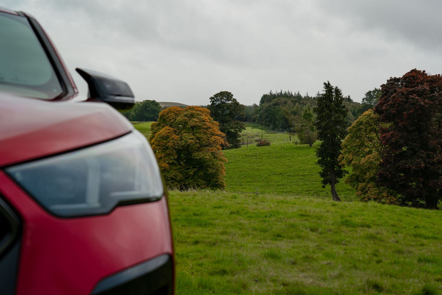 Red car driving through scenic countryside road with green rolling hills and autumn trees in the background.