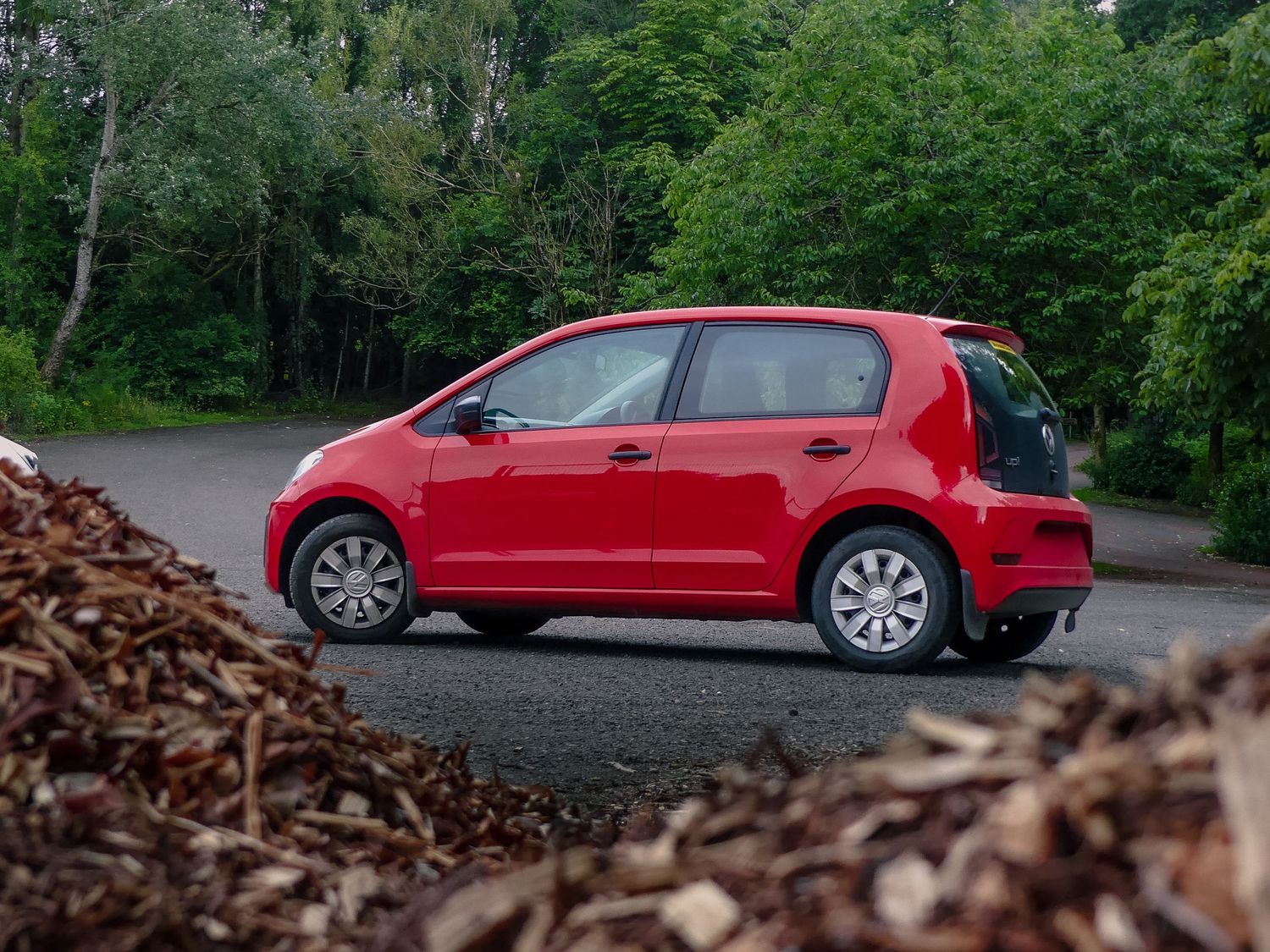 Red VW UP! parked on a gravel path surrounded by lush green forest scenery.