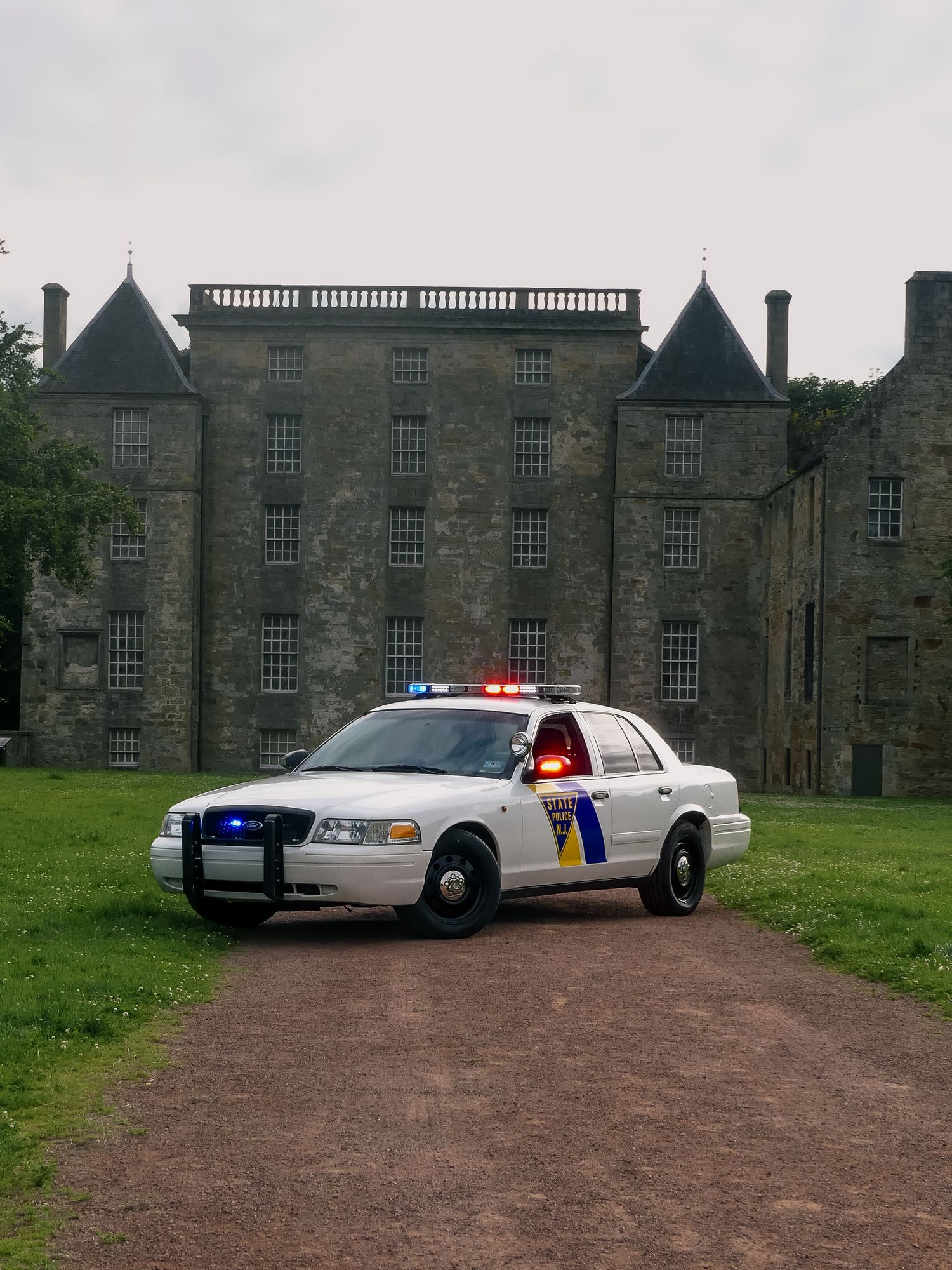 A police cruiser with flashing lights sits on a dirt path in front of a large stone manor house.