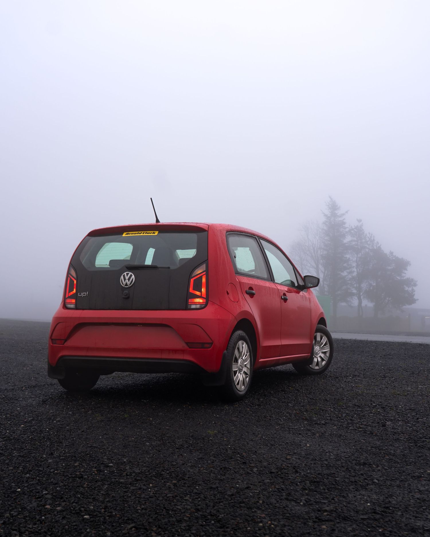 Red Volkswagen Up hatchback parked on dark gravel in misty foggy conditions with trees visible in background.