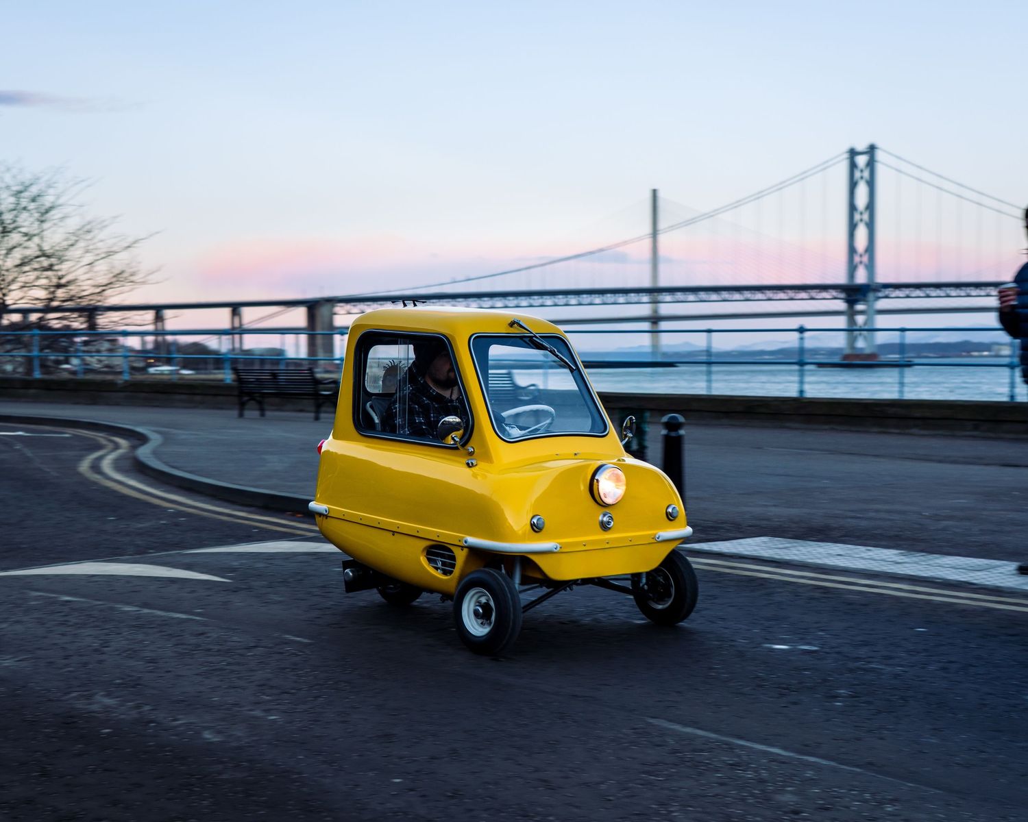 A bright yellow three-wheeled car drives along a waterfront road with a bridge visible in the background.