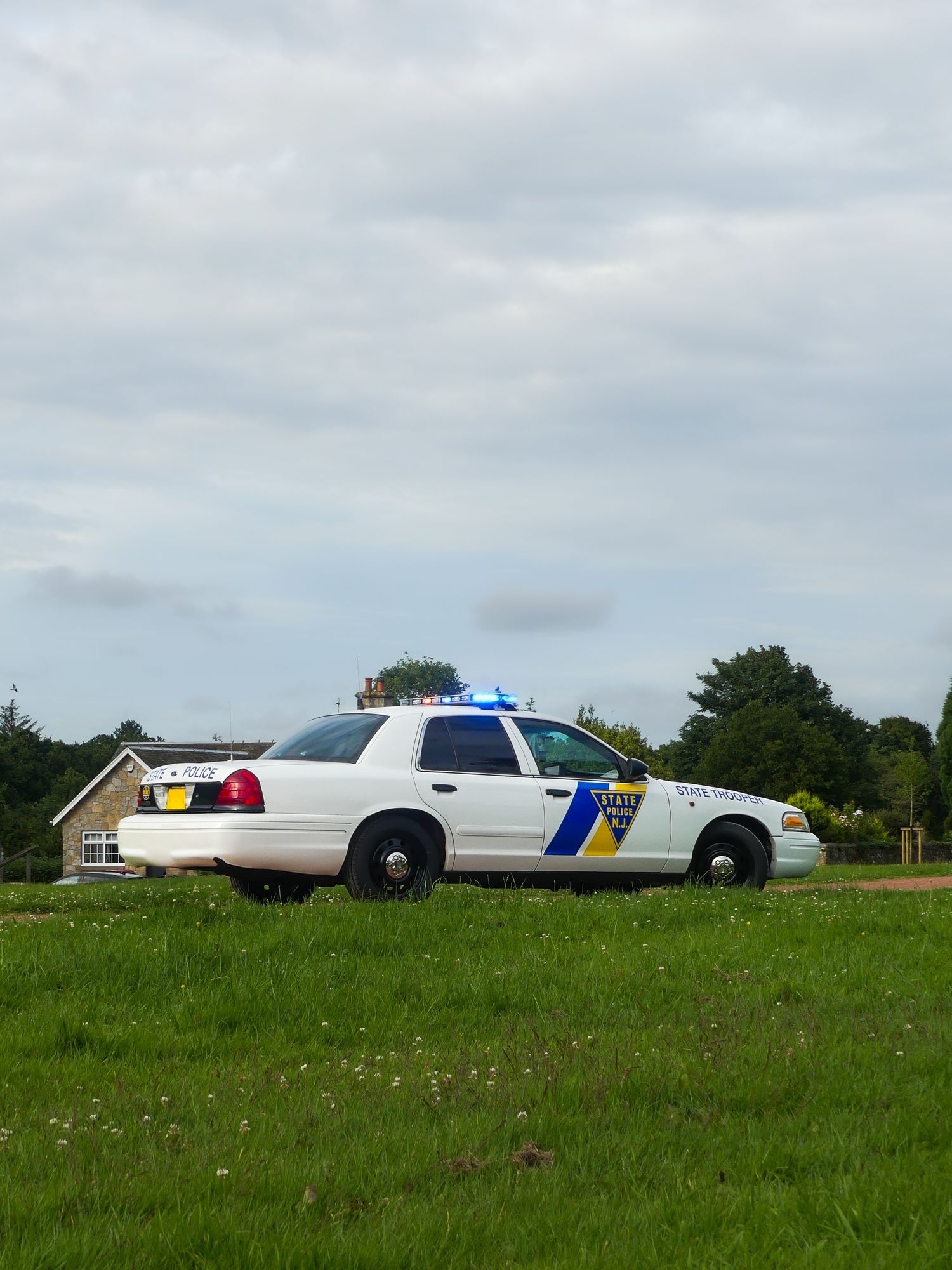 A white police patrol car with blue and yellow striping parked on grass with trees in the background.